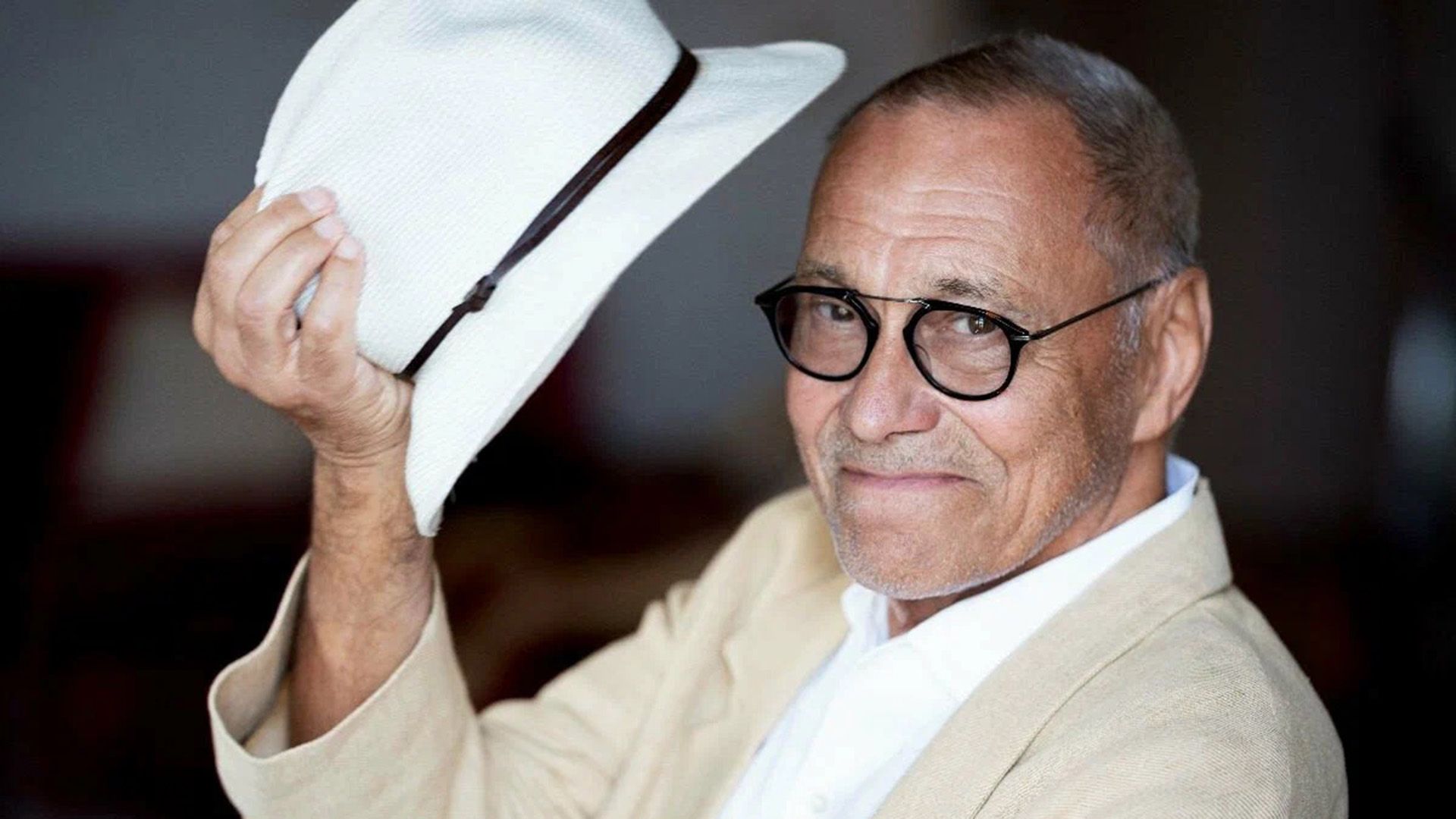 Un hombre de mediana edad con un traje claro sonríe y sostiene un sombrero blanco en la mano