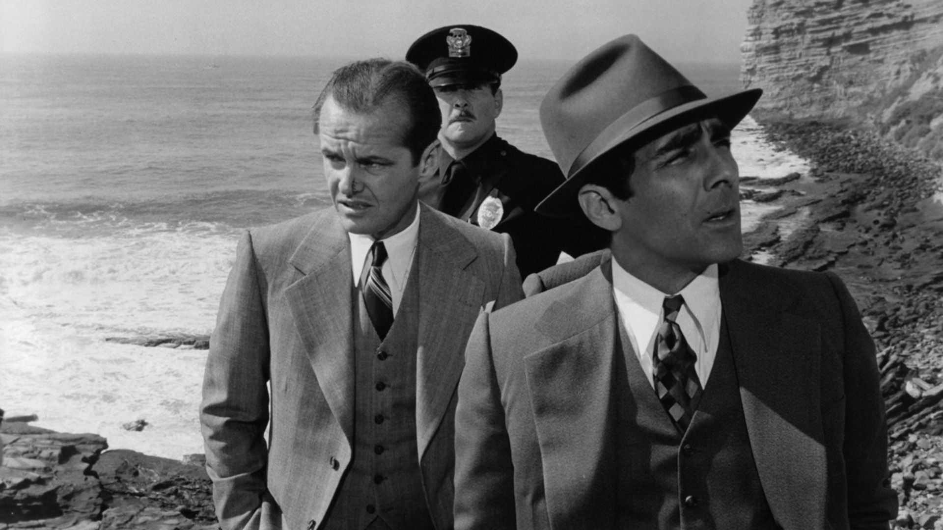 Una escena de una película en blanco y negro: dos hombres con trajes y un policía en uniforme están en la orilla junto a un acantilado