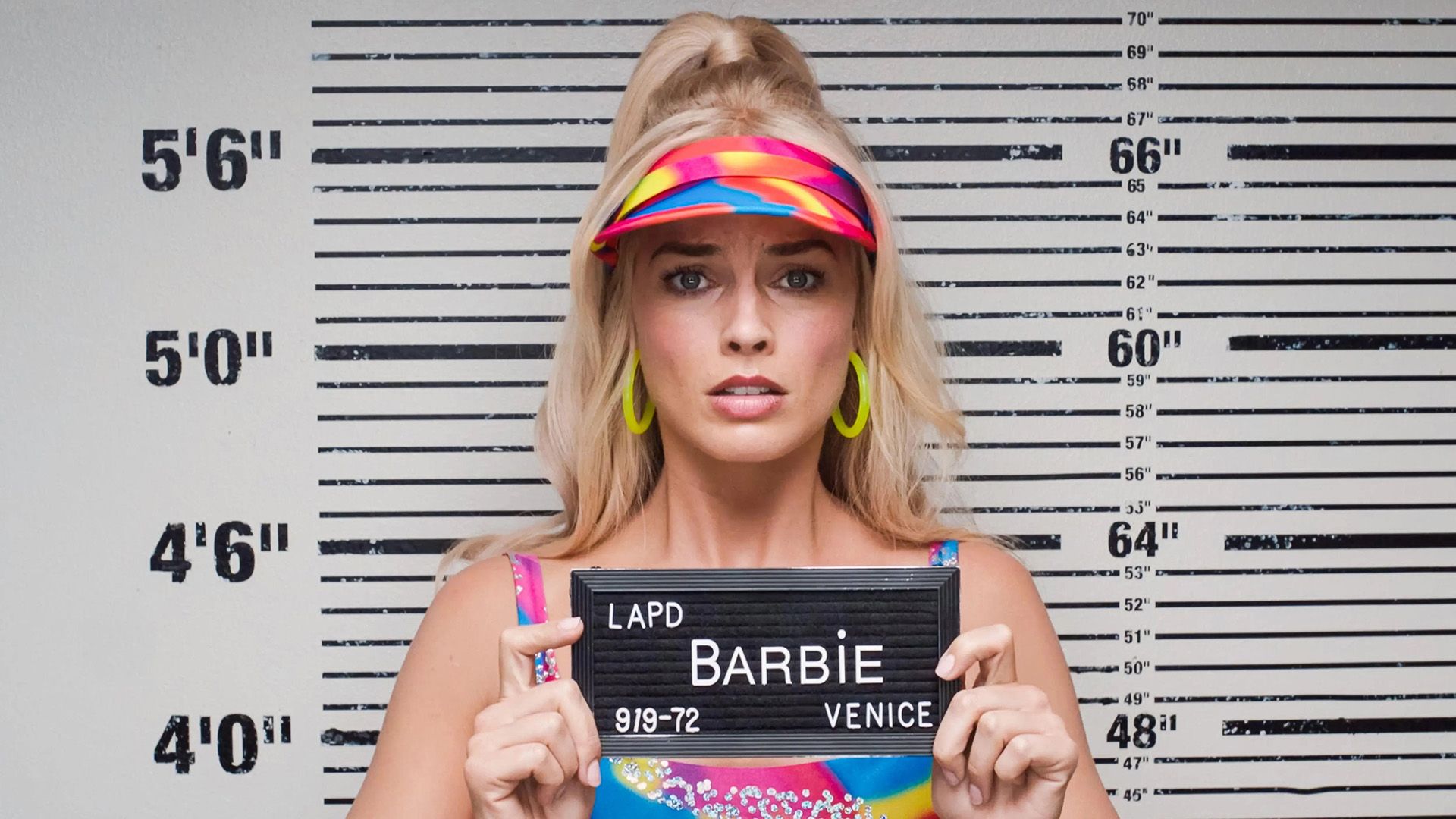 Foto policial: una rubia con una visera brillante se encuentra frente a una tabla de alturas sosteniendo un cartel con el nombre "barbie"