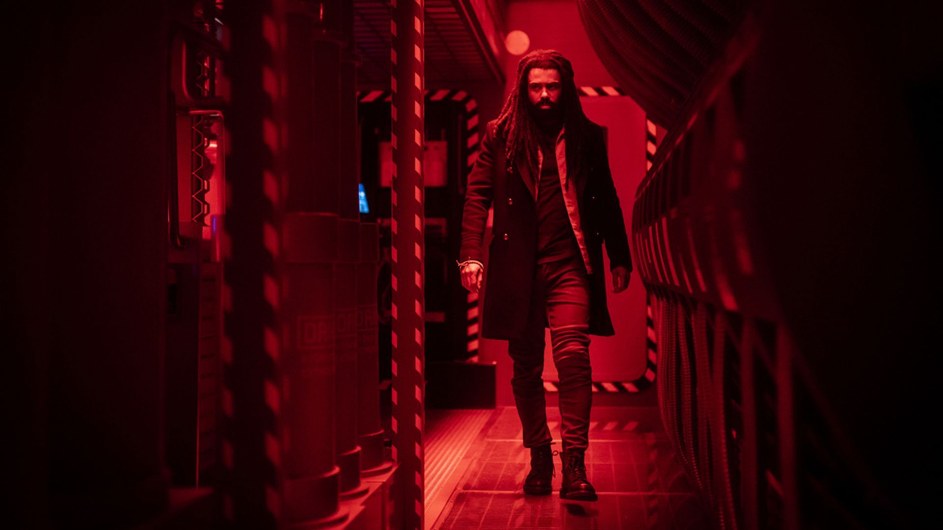 A man with dreadlocks walks down a corridor bathed in red light