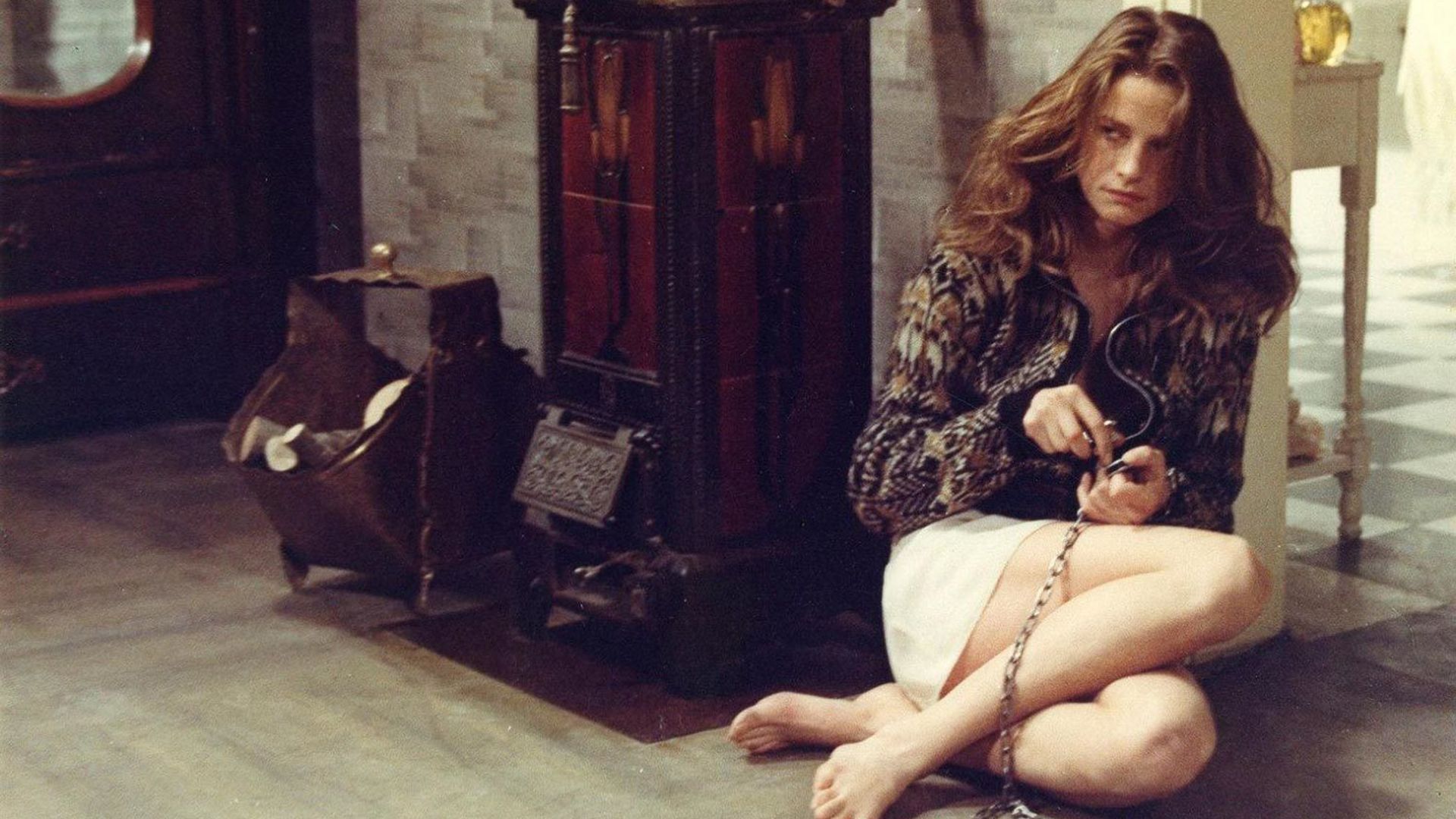 Joven Charlotte Rampling con cabello largo sentada en el suelo junto a la estufa, encadenada, intentando liberarse.