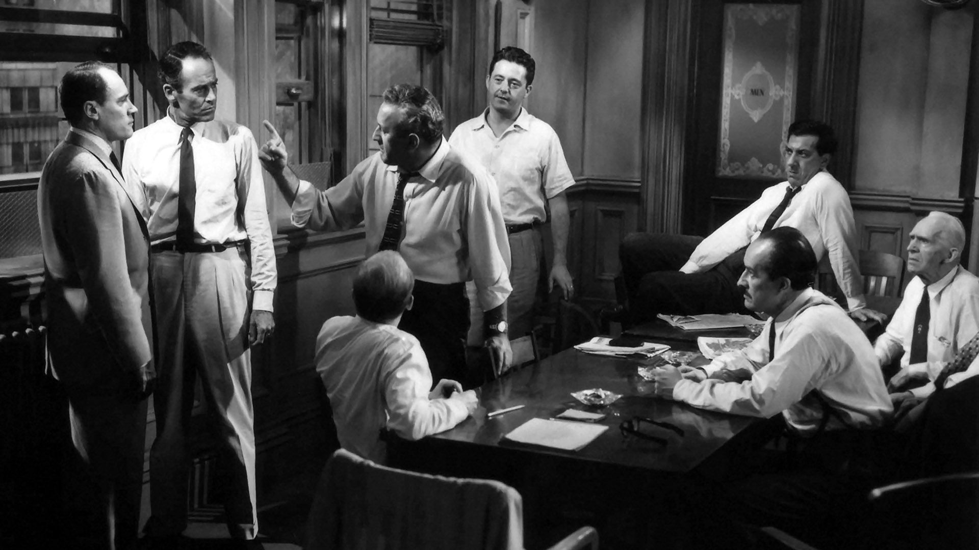 Doce hombres en una habitación, algunos sentados en la mesa, otros de pie, discutiendo