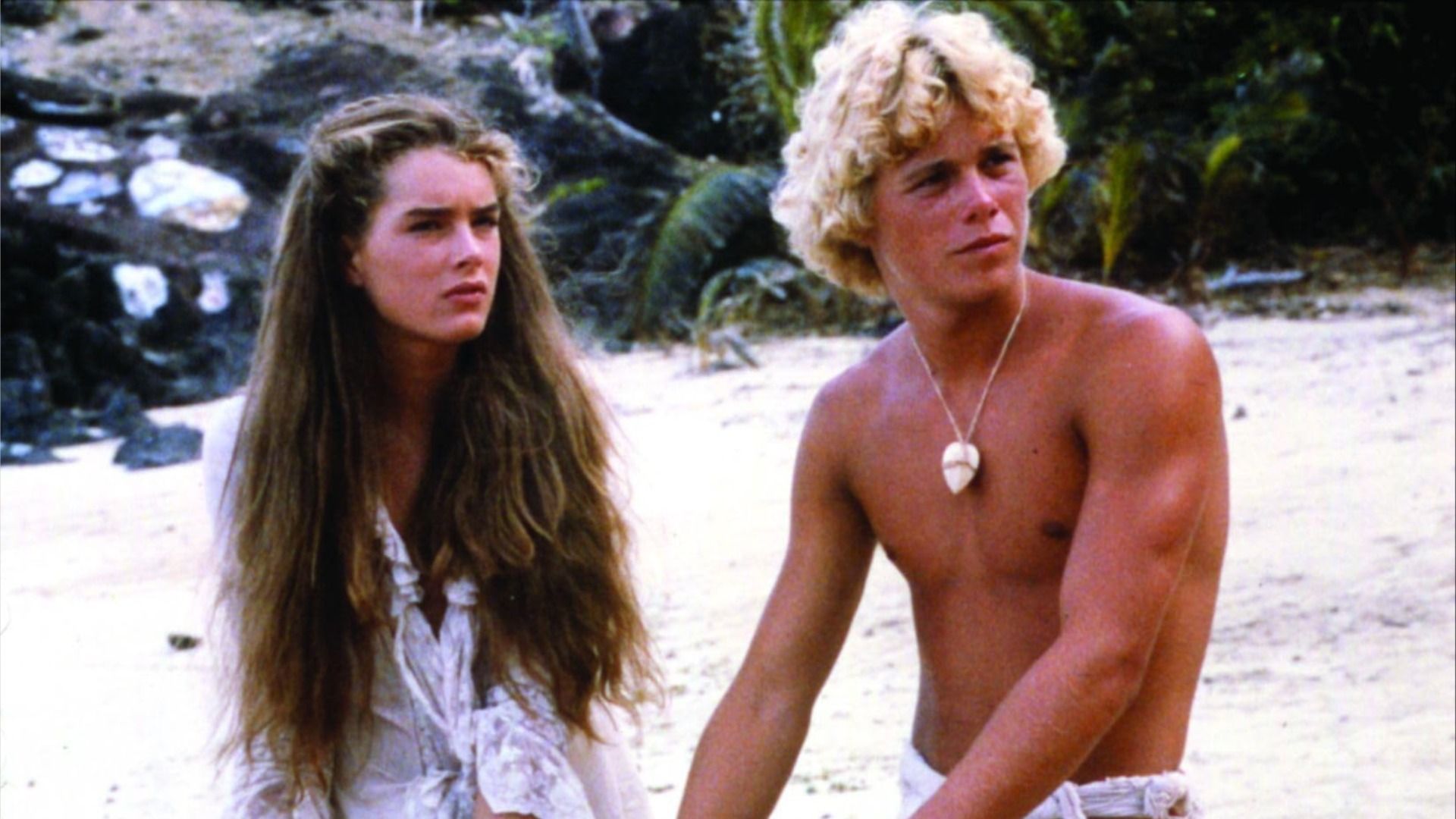 Los actores Brooke Shields y Christopher Atkins sentados en la playa con ropa hecha jirones