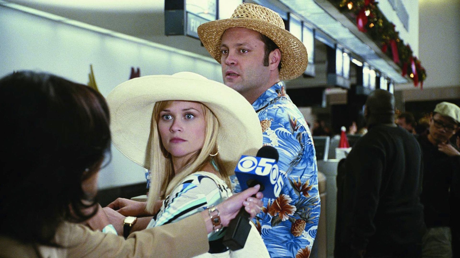 Aeropuerto, Reese Witherspoon y Vince Vaughn con sombreros de verano se ven sorprendidos ante una periodista con un micrófono en la mano
