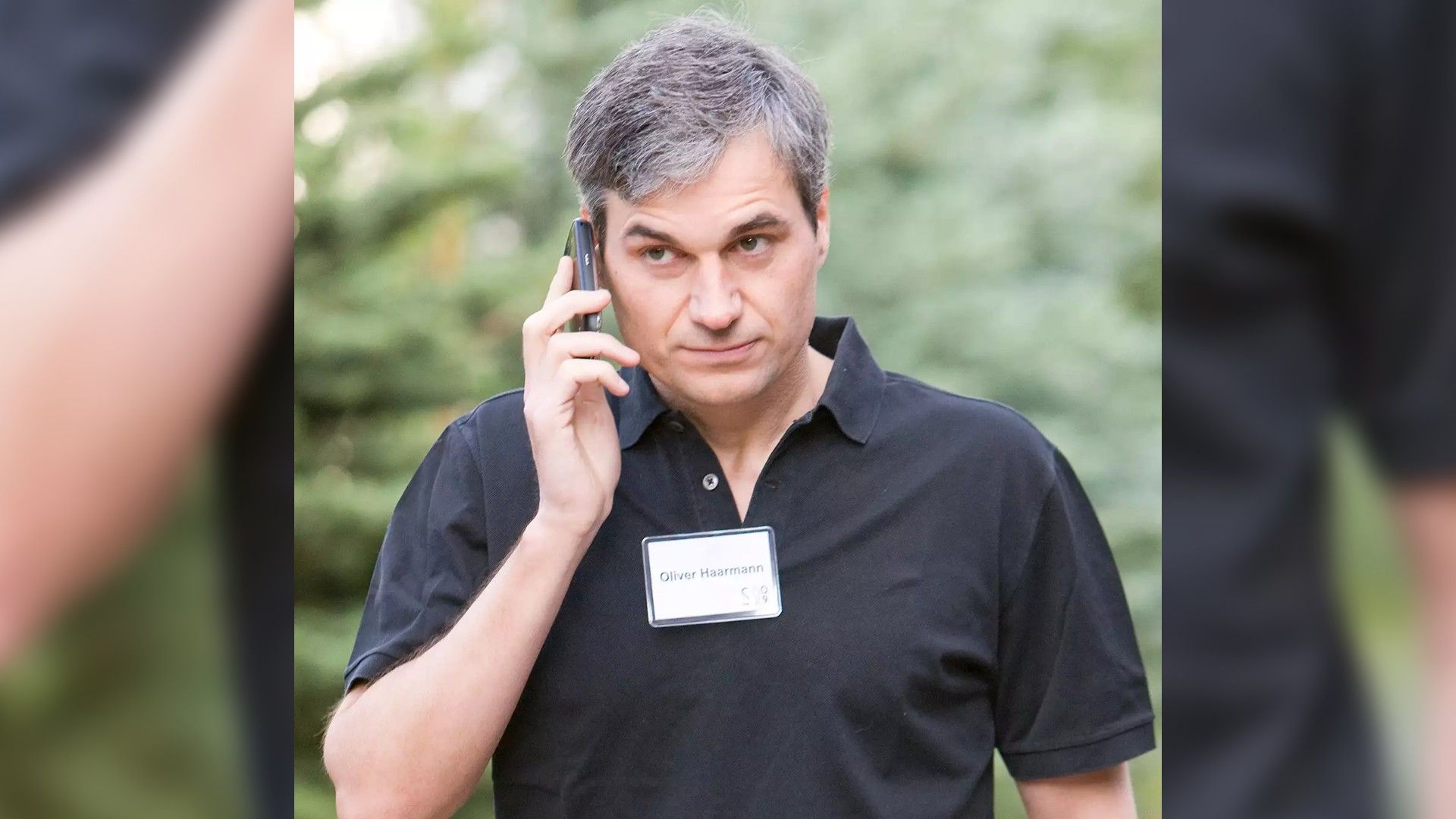 Un hombre con camisa negra y una placa hablando por teléfono contra un fondo de árboles verdes, con aspecto concentrado