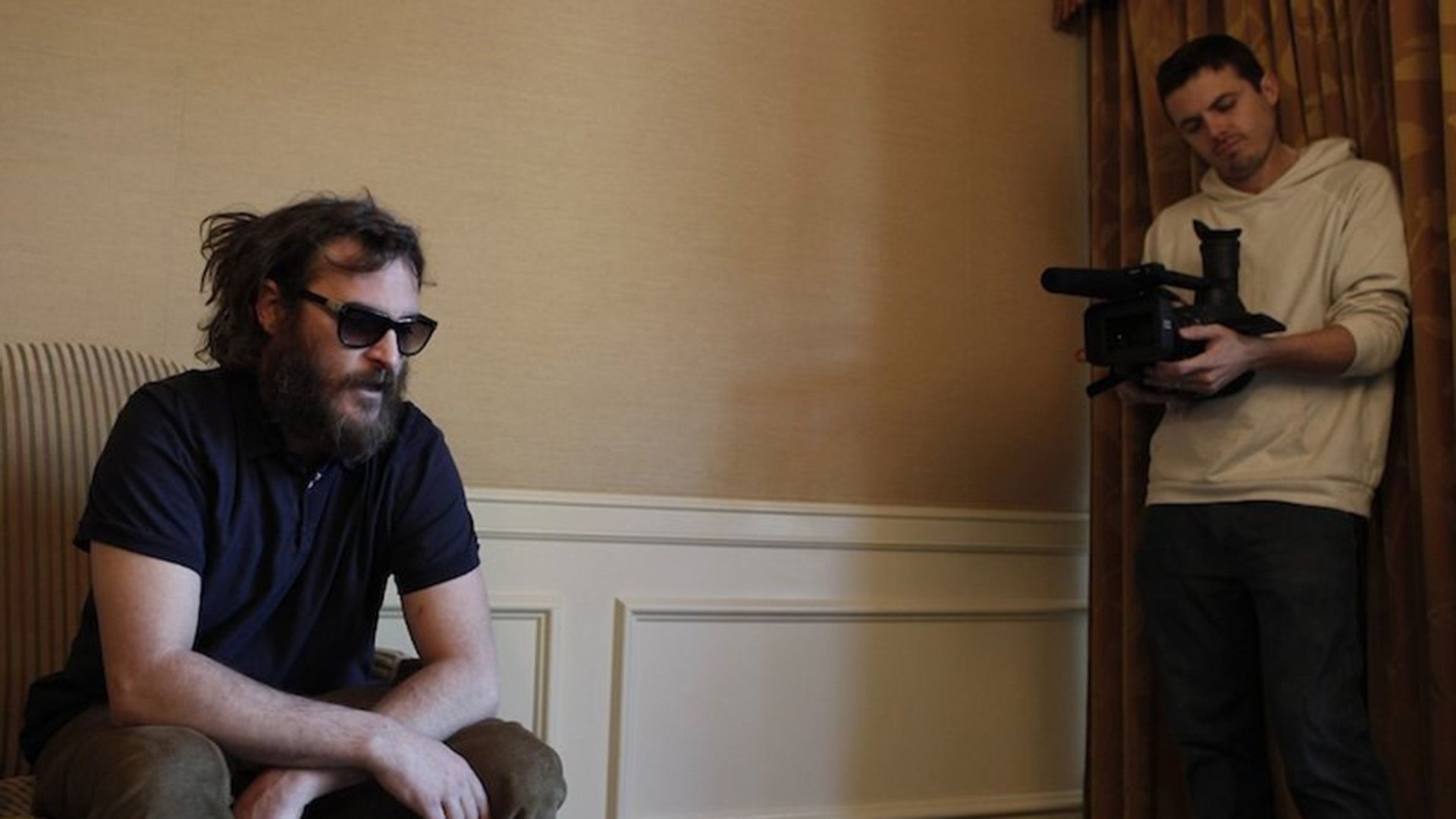 Casey Affleck con una cámara en mano filmando a Joaquin Phoenix sentado en una silla con gafas oscuras