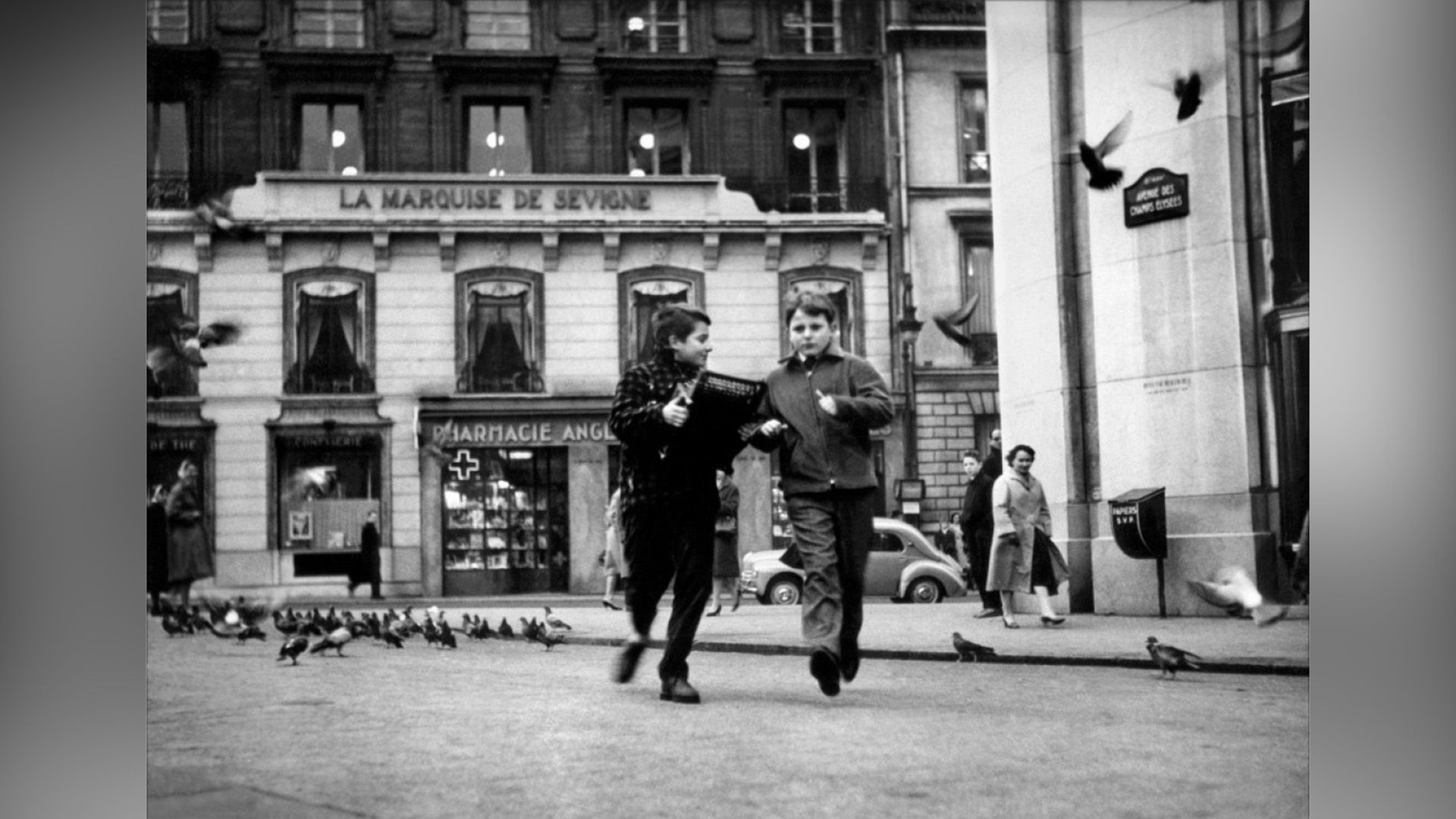Dos chicos corren por una calle de París, rodeados de transeúntes y palomas