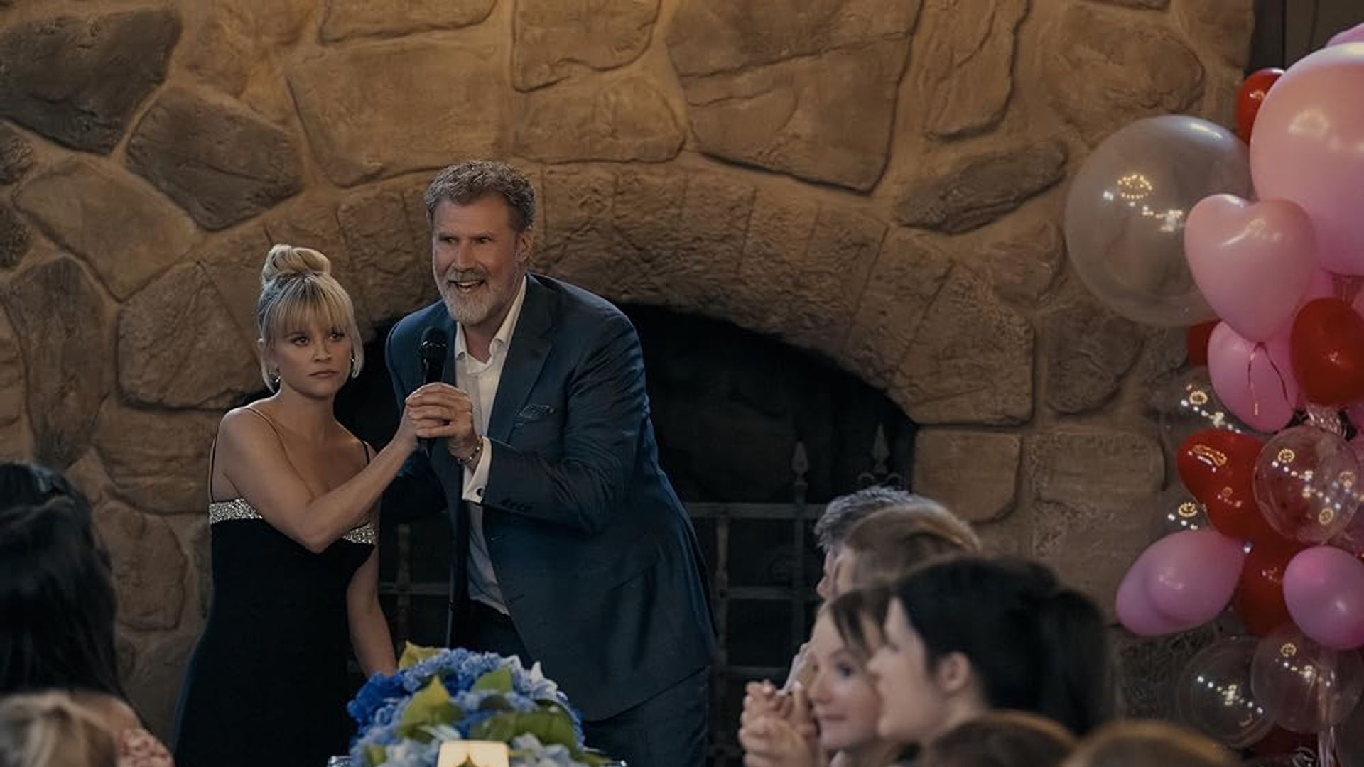 Will Ferrell con traje y micrófono toma de la mano a Reese Witherspoon en vestido negro contra el fondo de una chimenea, mesa festiva y globos