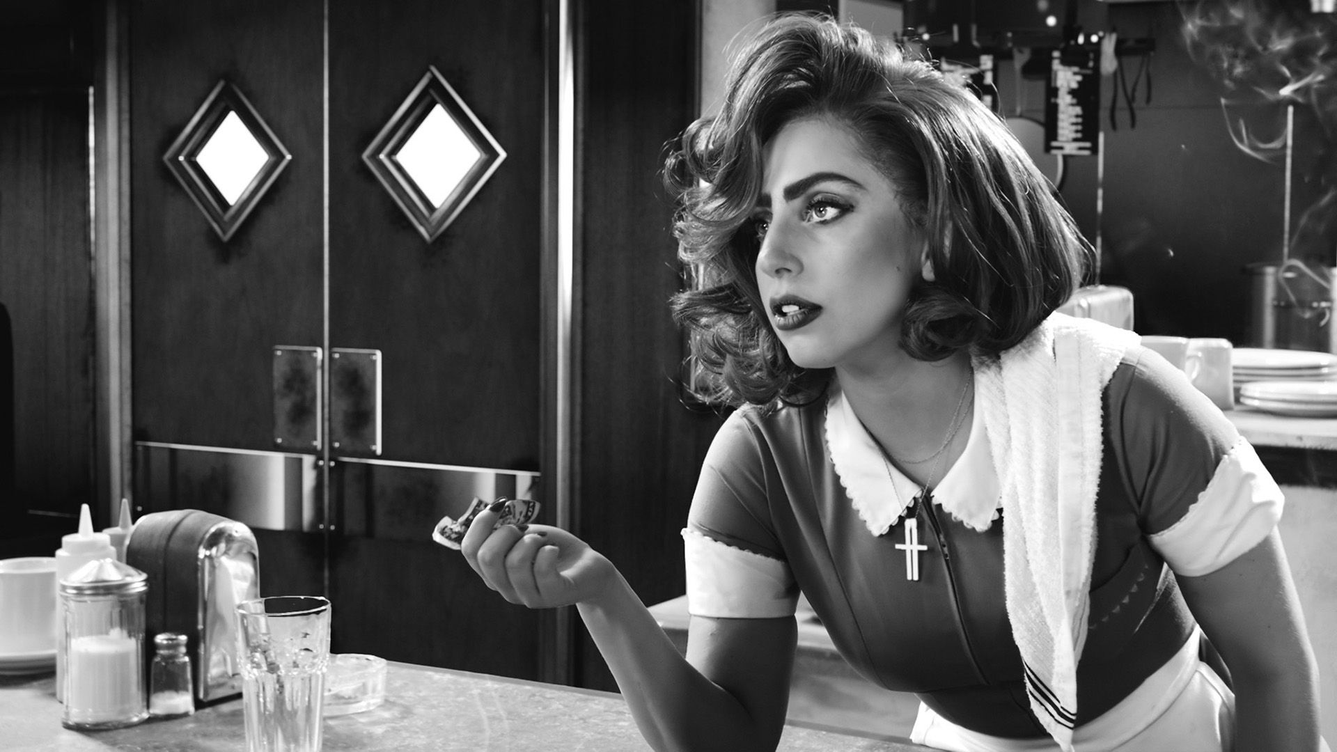 Lady Gaga en un atuendo de camarera apoyada en el mostrador del bar, sosteniendo una cuenta en su mano