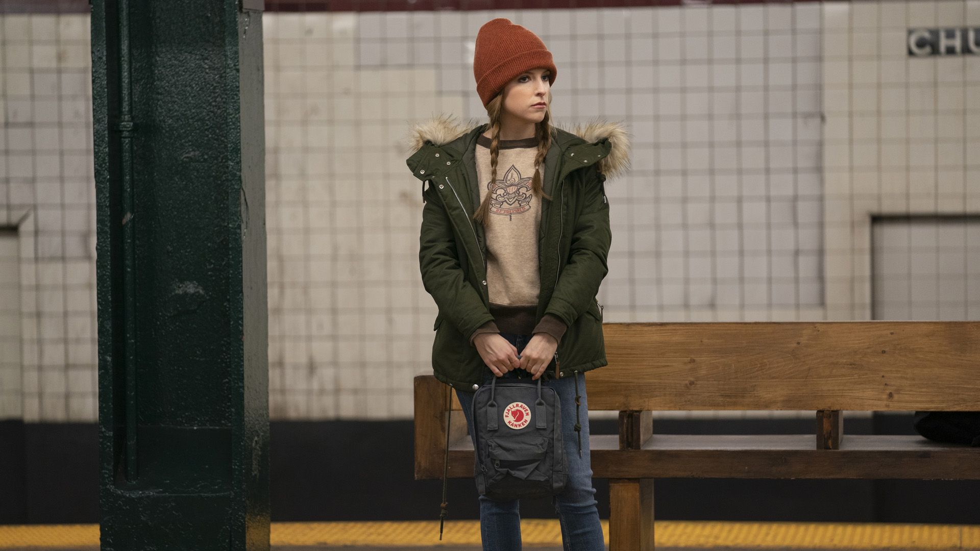 Anna Kendrick con un sombrero rojo y una parka verde en la estación de metro