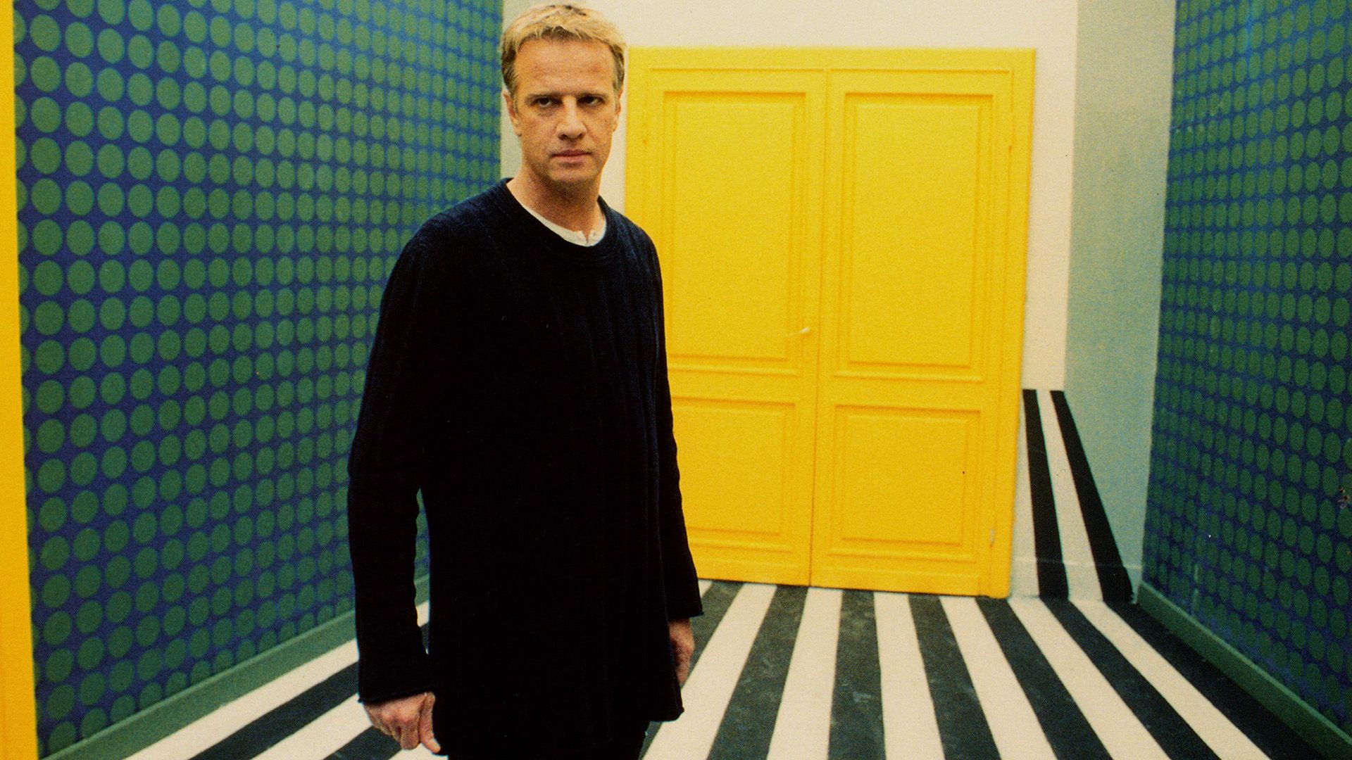 Christopher Lambert está en una habitación con paredes verdes, un piso en blanco y negro, y una puerta amarilla