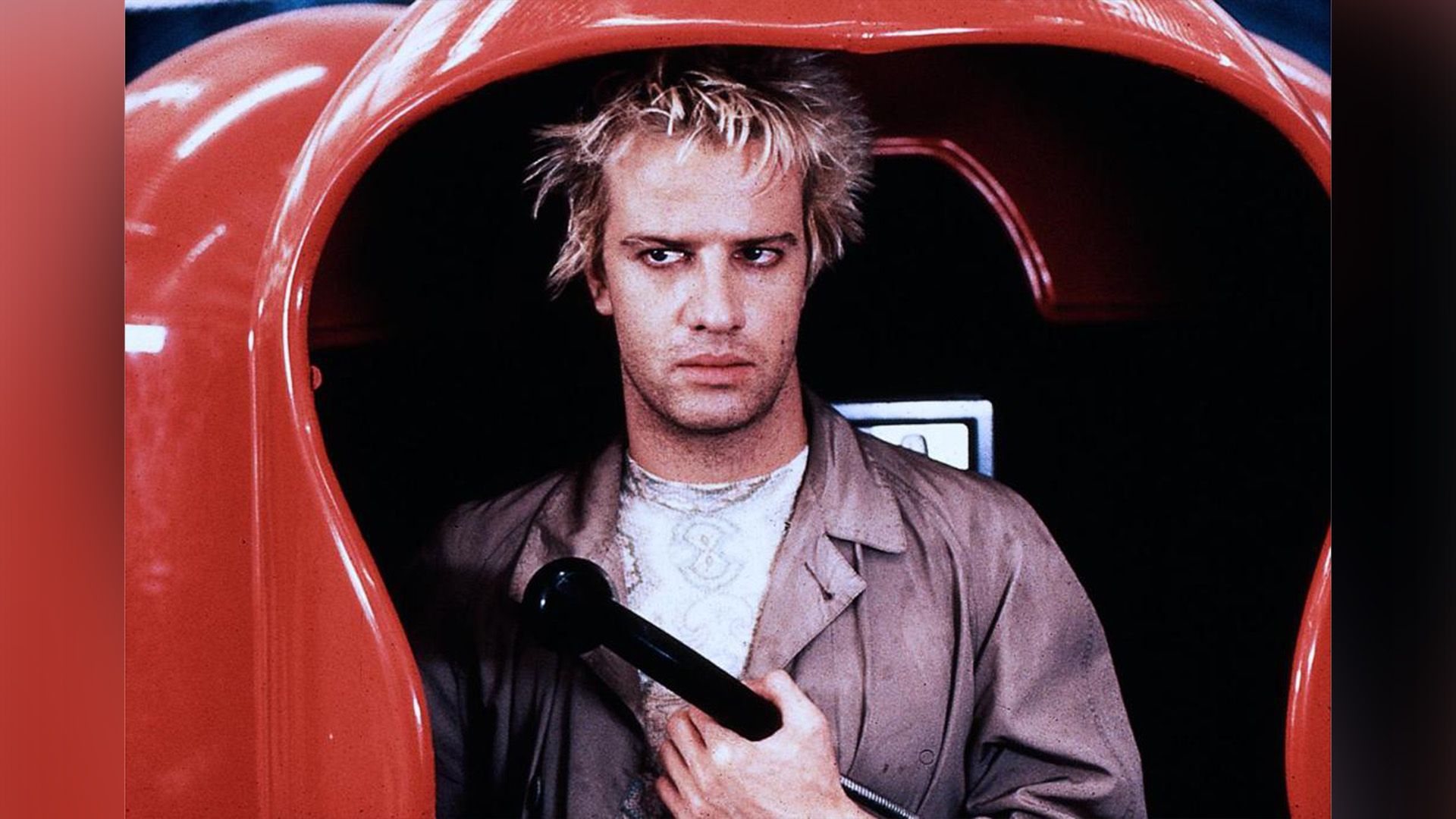 Joven Christopher Lambert con cabello blanco de pie en una cabina telefónica roja