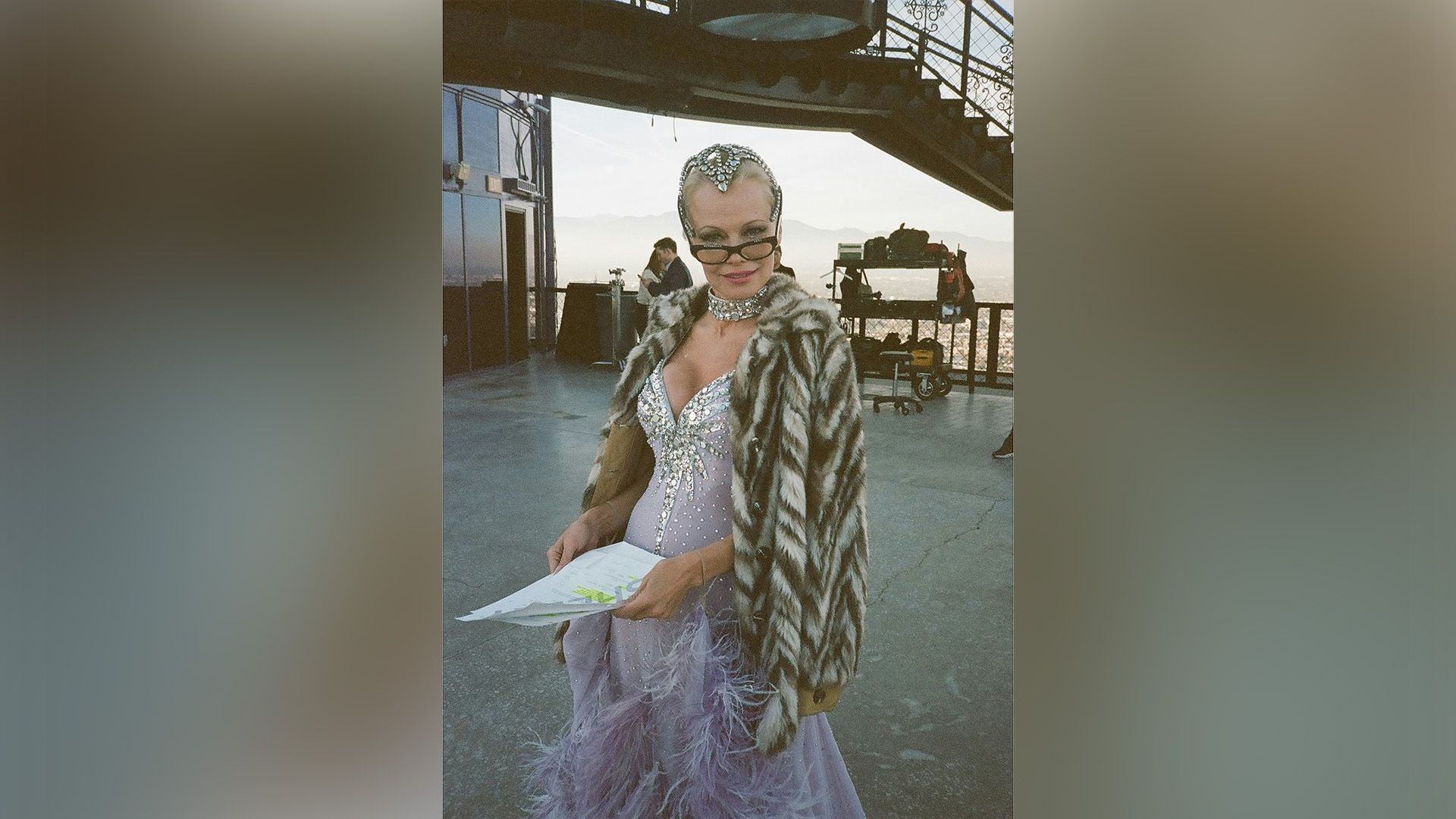 Pamela Anderson en un vestido lila con pedrería y plumas sostiene un guion en sus manos y mira a la cámara por encima de sus gafas