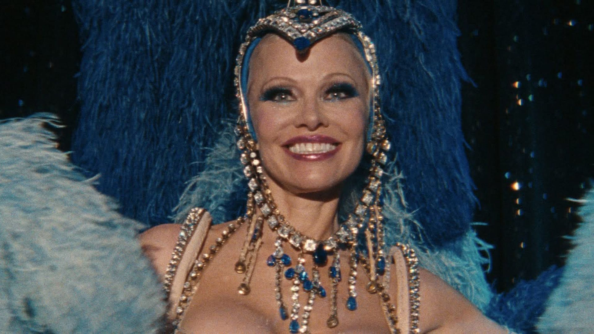 Pamela Anderson con un atuendo de plumas azules y turquesas, cuentas y pedrería