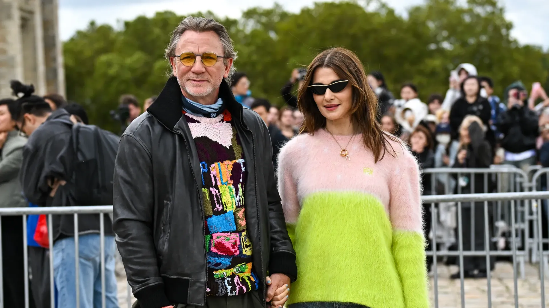 Rachel Weisz con un suéter blanco y verde y Daniel Craig con una chaqueta de cuero negra y un suéter colorido tomados de la mano
