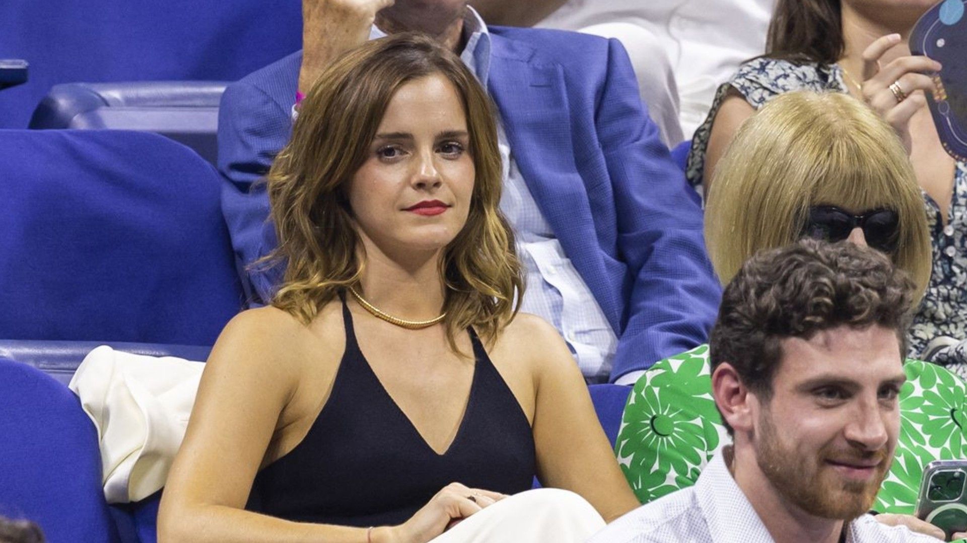 Emma Watson en las gradas de un evento deportivo, mirando hacia adelante, sentada con un top negro y un collar dorado