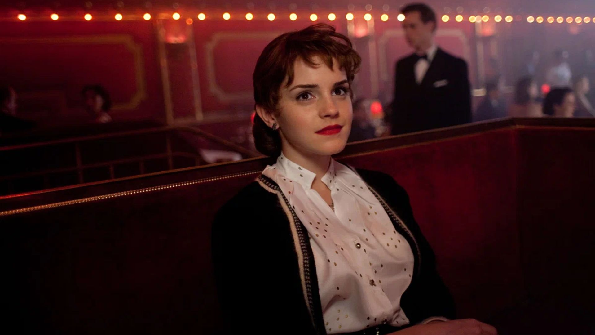 Emma Watson con un peinado y lápiz labial rojo sentada en un club oscuro