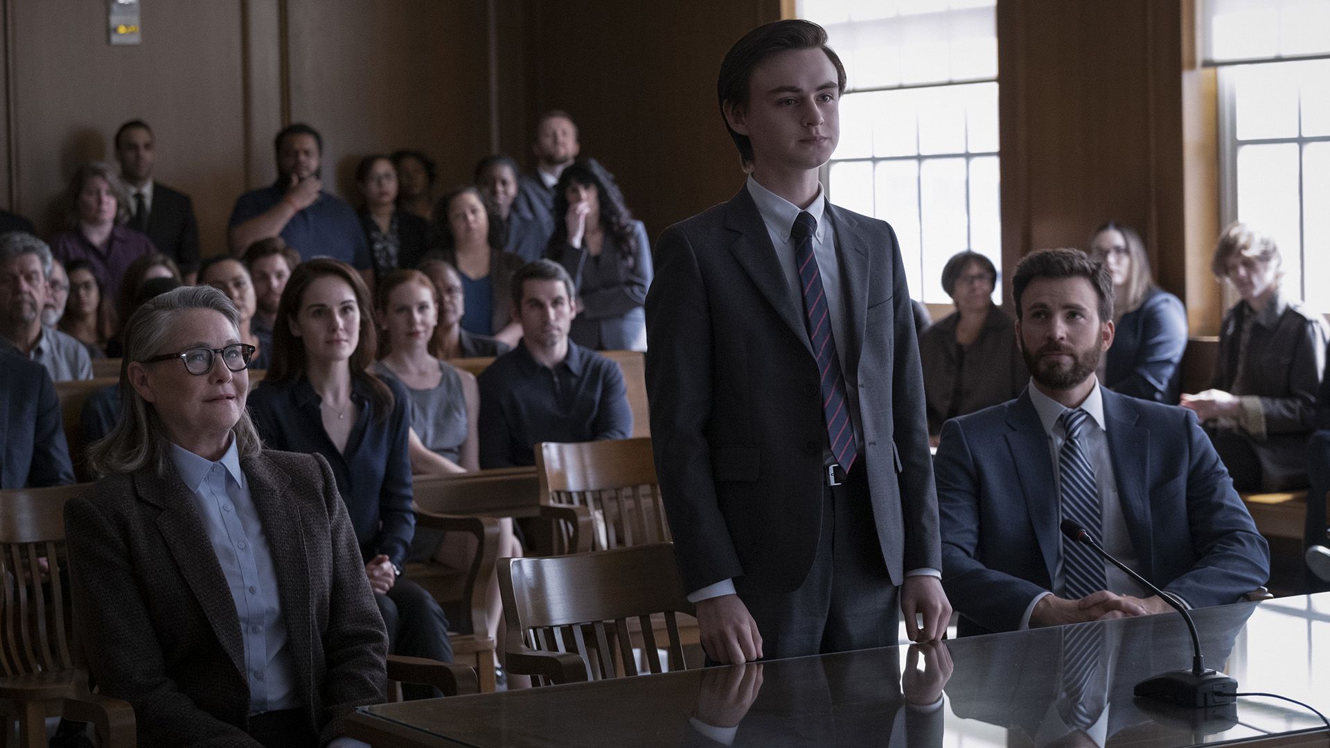 Jaeden Martell está de pie en la mesa del tribunal, sentado junto a él está Chris Evans, con espectadores al fondo
