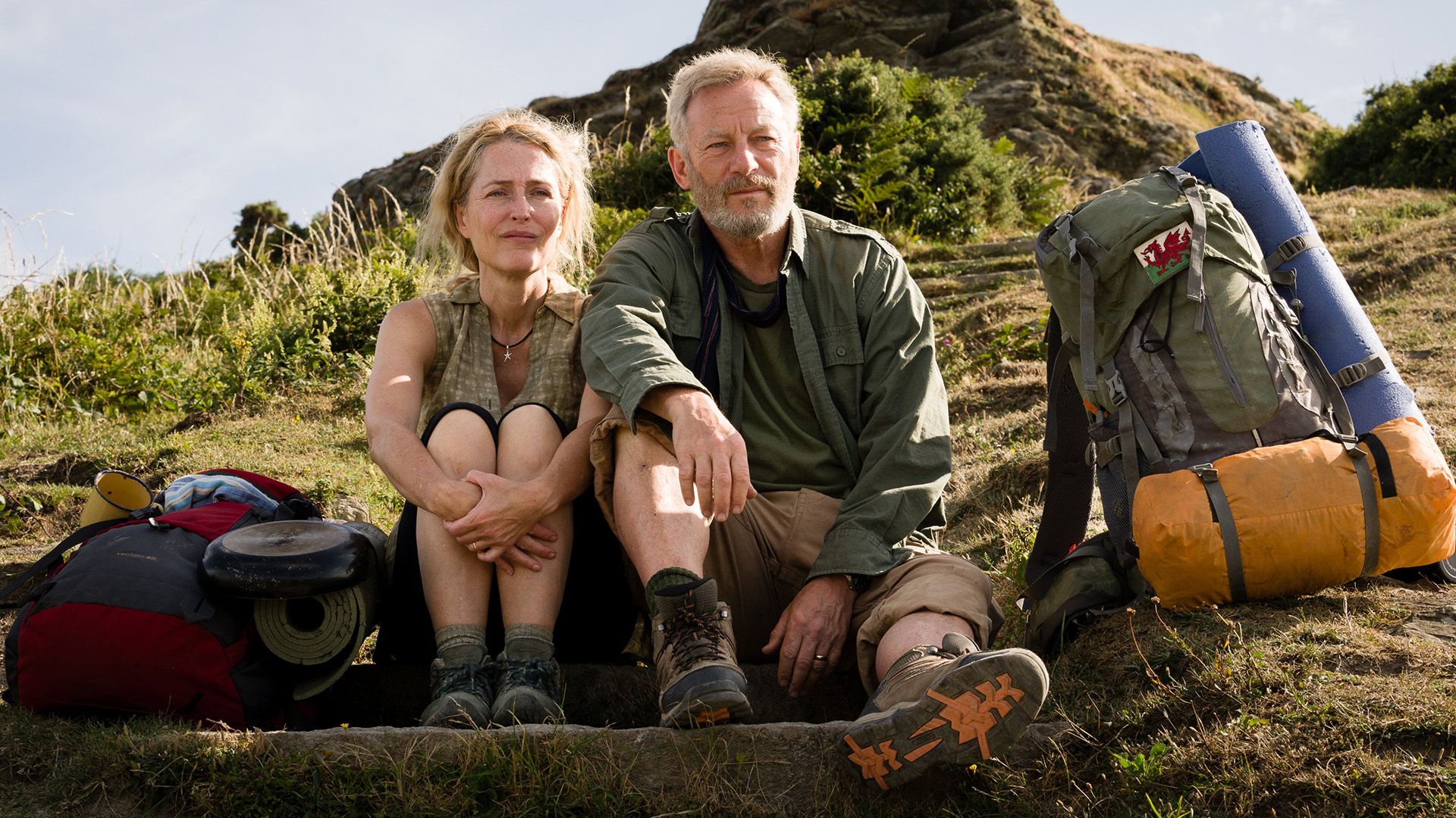 Gillian Anderson y Jason Isaacs están sentados en una colina, sus mochilas de senderismo están junto a ellos