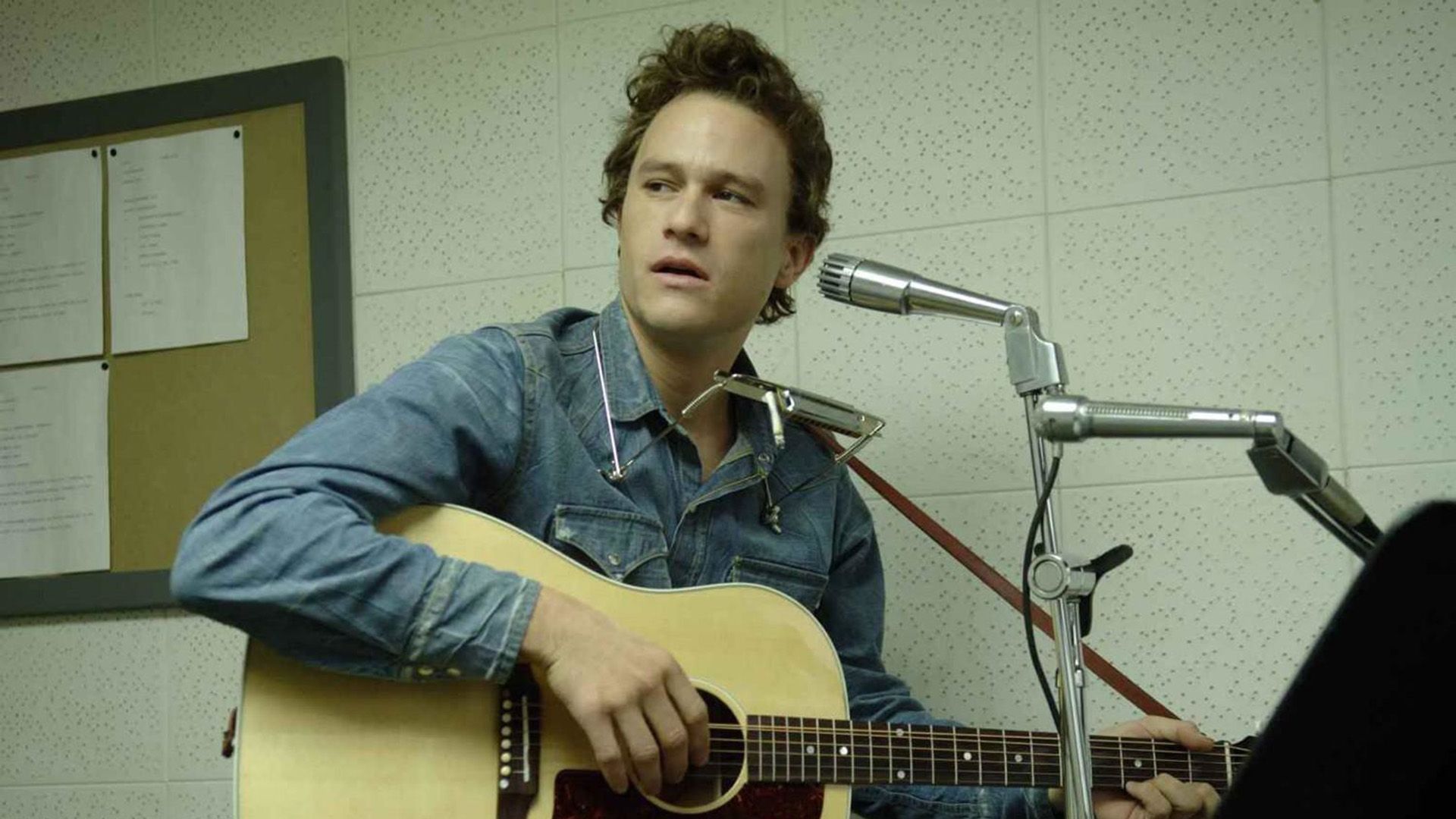Heath Ledger con una guitarra y armónica frente a un micrófono en la película "I'm Not There"