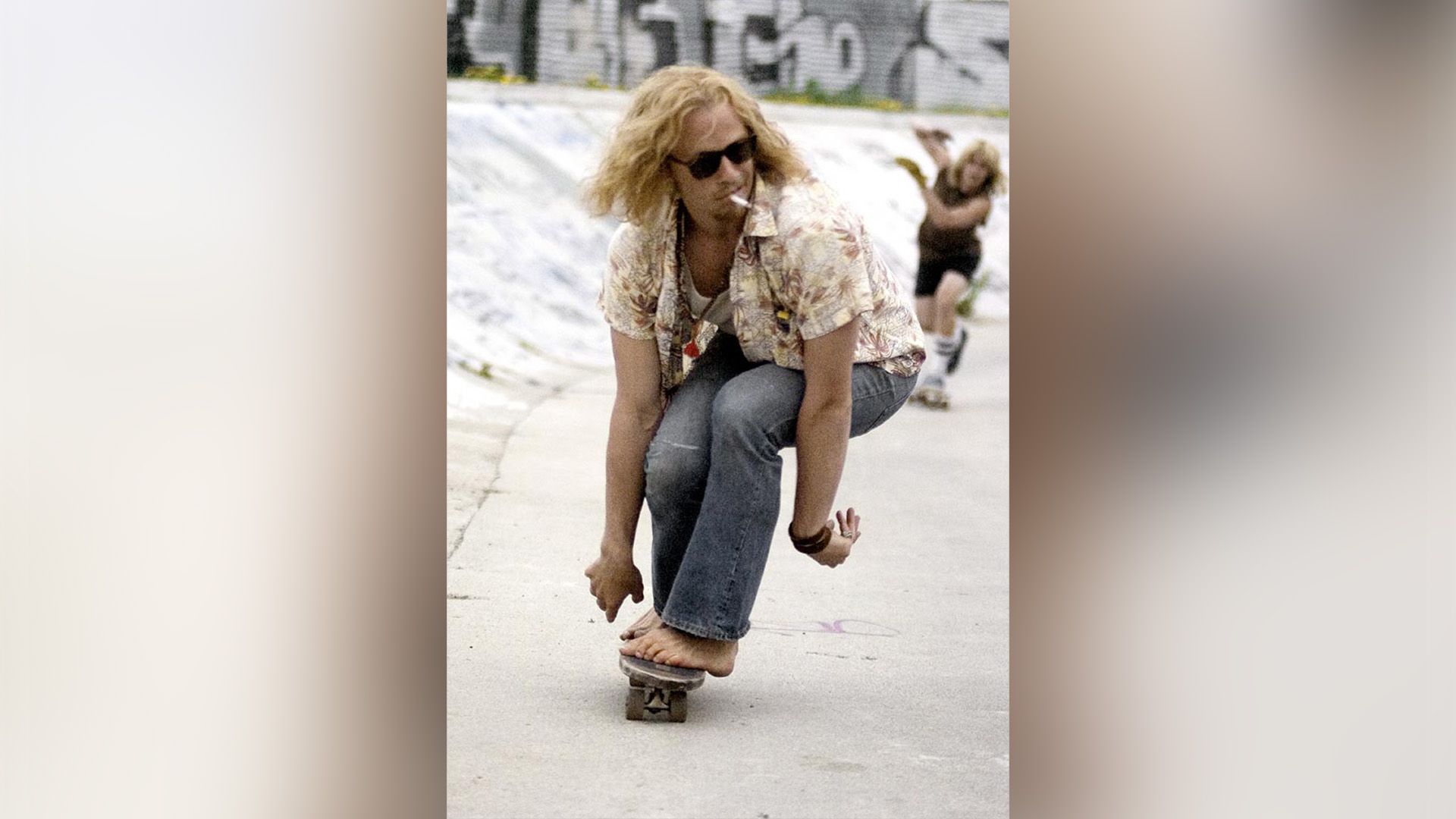 Heath Ledger con el pelo largo y gafas oscuras montando una patineta descalzo