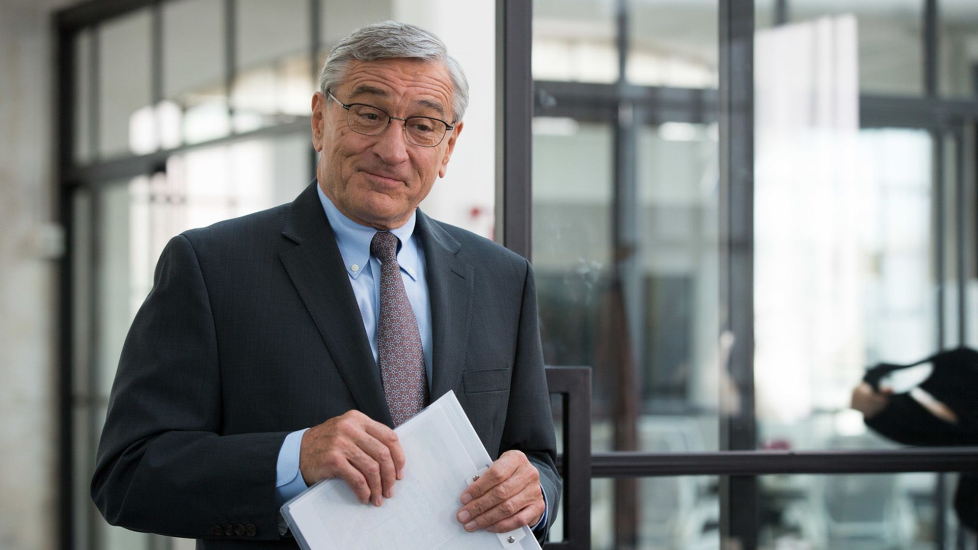 Robert De Niro con una chaqueta, corbata y gafas sostiene una carpeta para papeles en sus manos