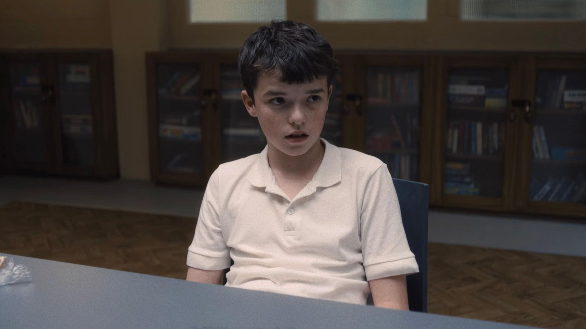 El actor Owen Cooper con una camiseta blanca sentado en una mesa, con estanterías de libros al fondo
