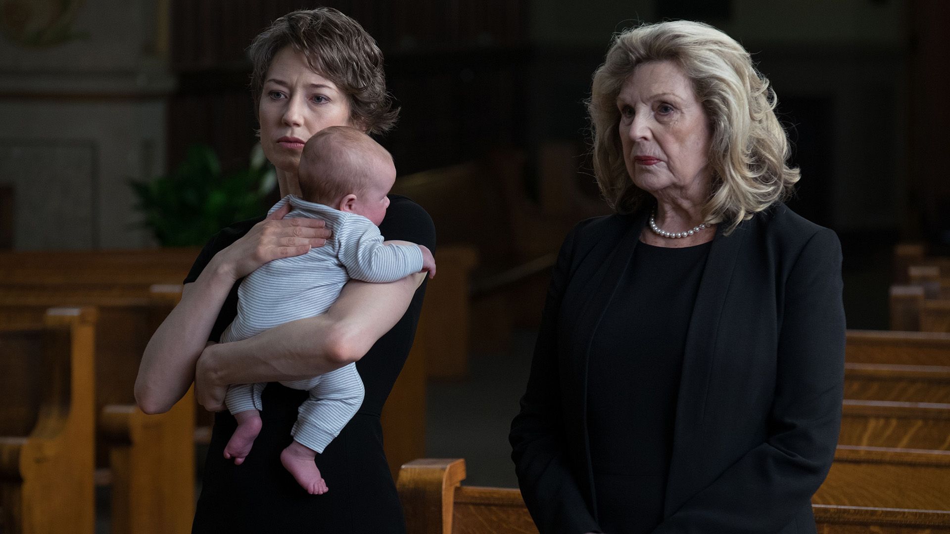 Carrie Coon con un bebé en brazos y una mujer mayor vestida de negro de pie en la iglesia