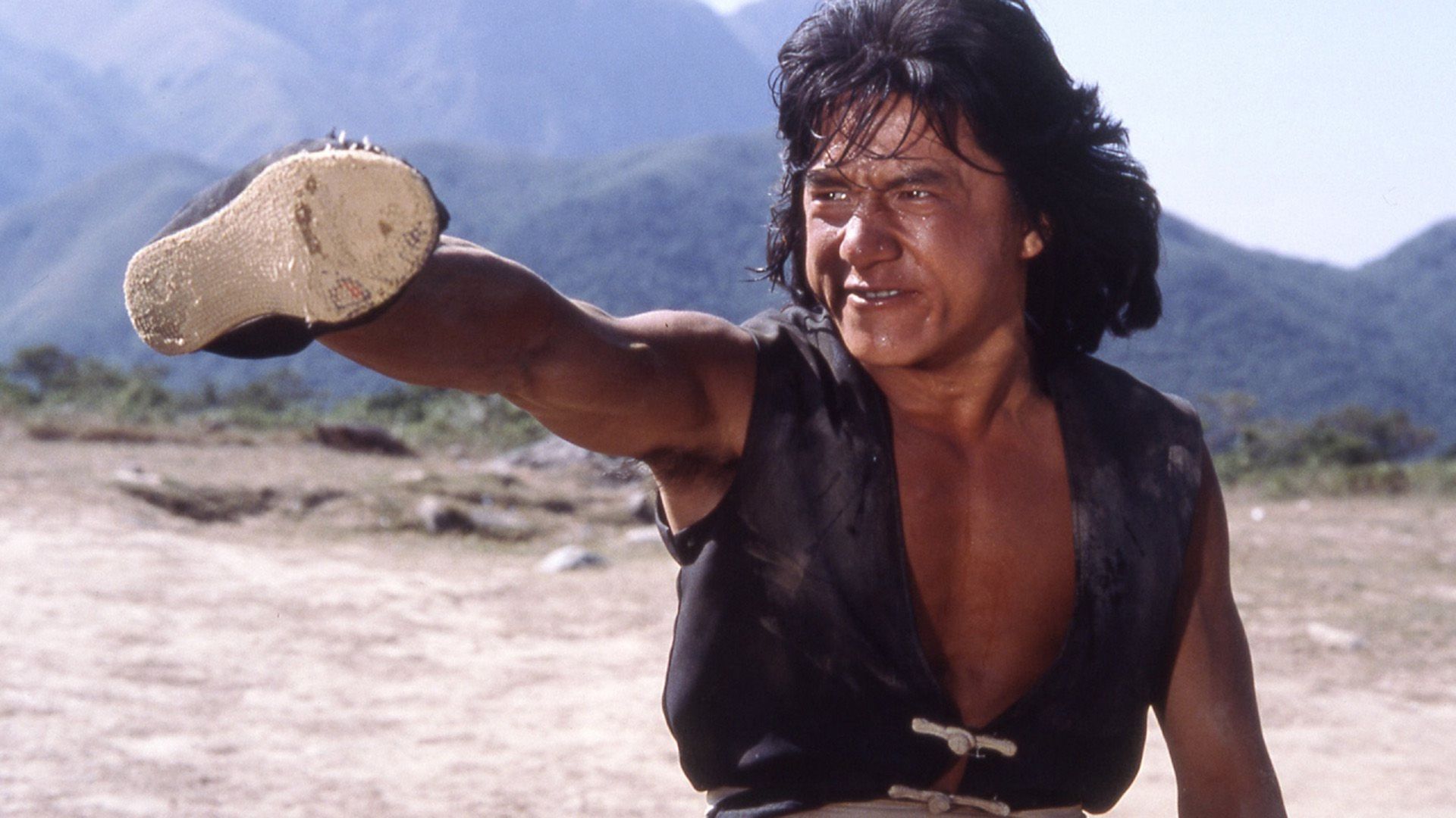 Joven Jackie Chan en un chaleco negro lanzó su brazo hacia adelante con un zapato en un golpe