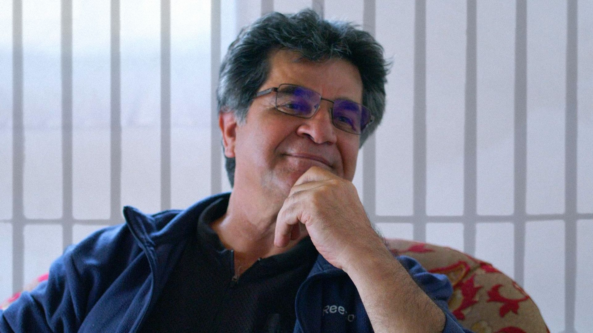 Director Jafar Panahi in a blue jacket sits in a chair and smiles, looking to the side