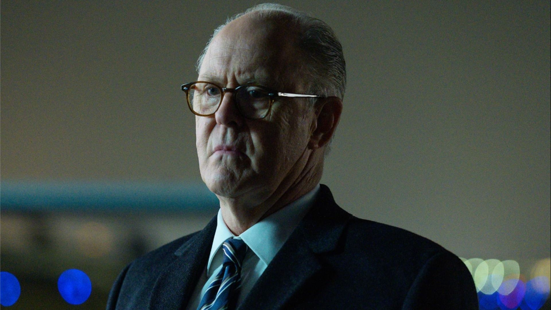 Actor John Lithgow en un traje y gafas, parado sin expresión y mirando hacia un lado