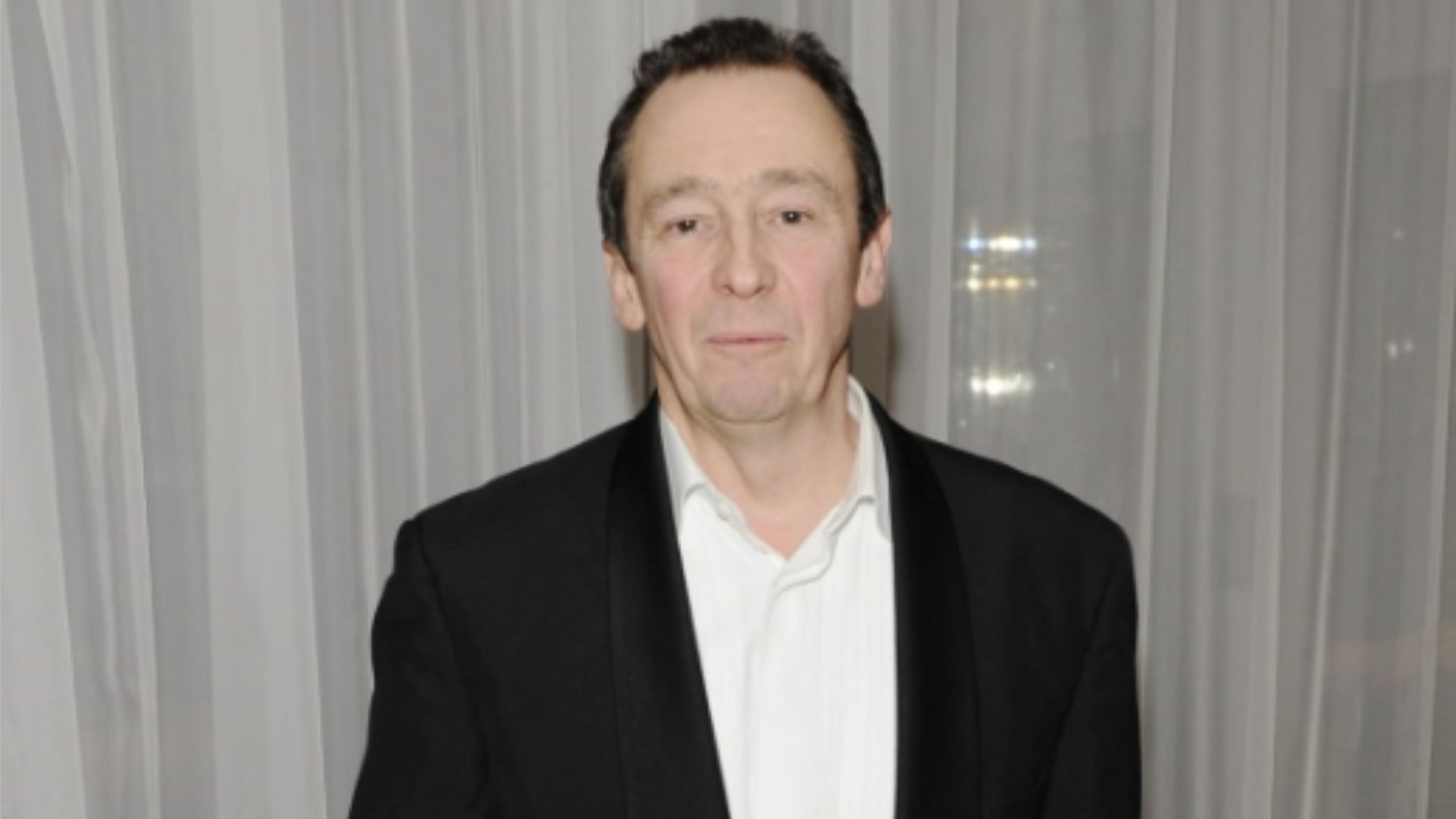 Actor Paul Whitehouse en un traje negro contra un fondo de tul blanco, mirando a la cámara
