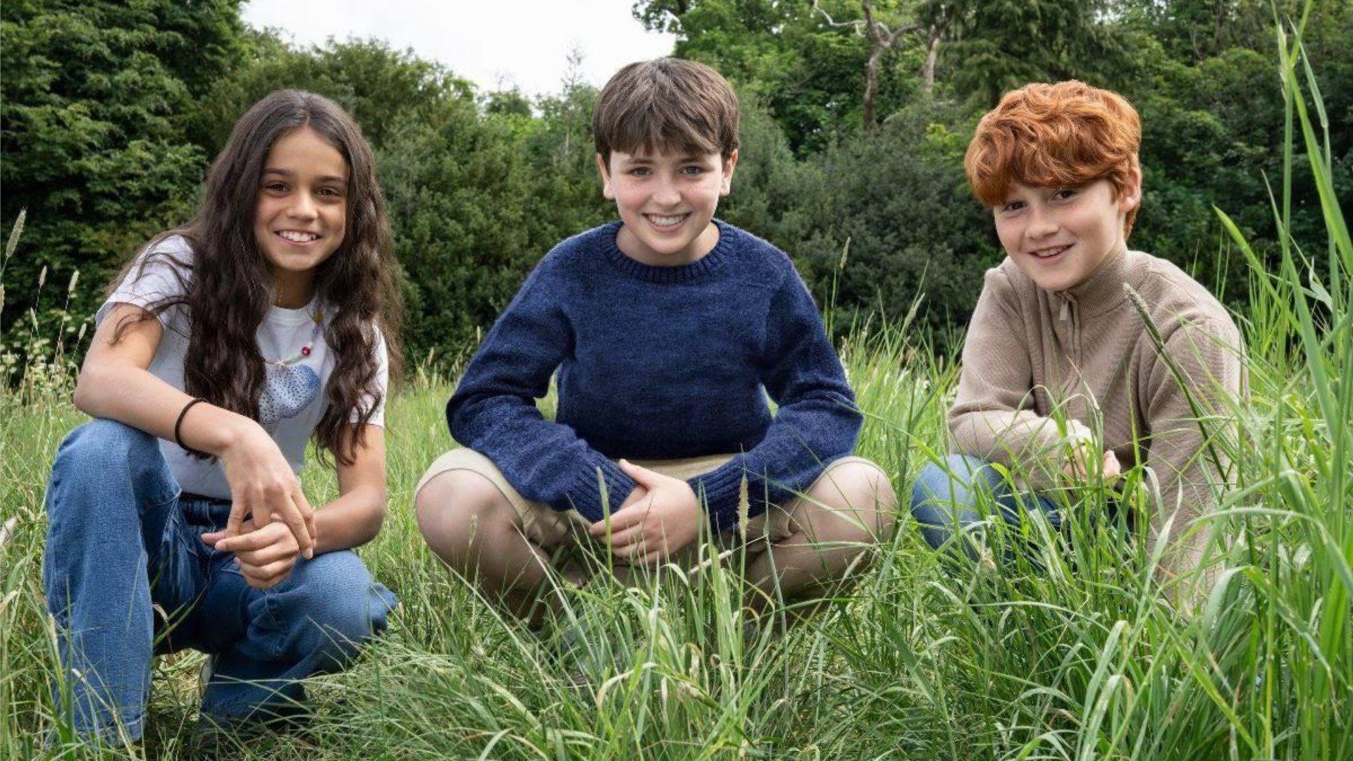 Actores Dominic McLaughlin, Alastair Stout y Arabella Stanton sentados en el césped y sonriendo