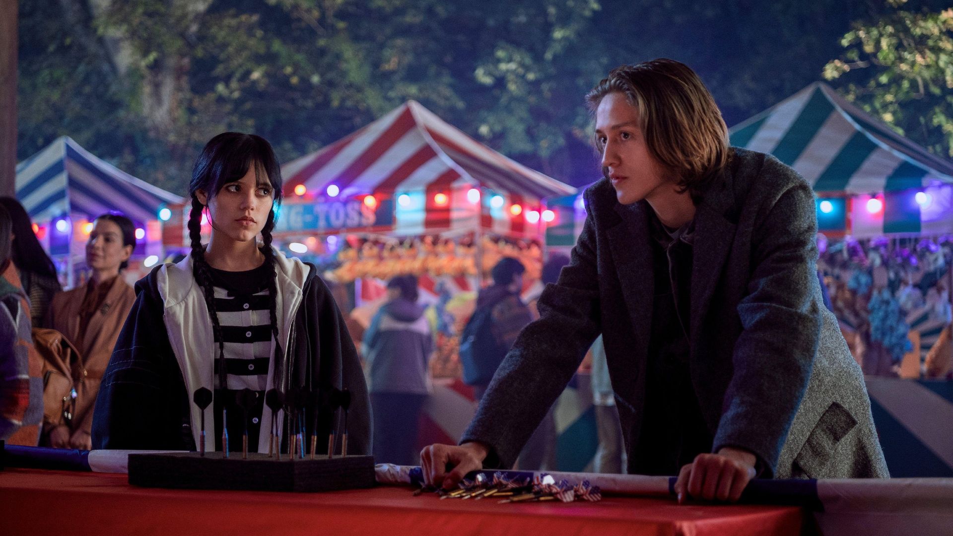 Jenna Ortega y Percy Hynes White en primer plano con una feria de fondo