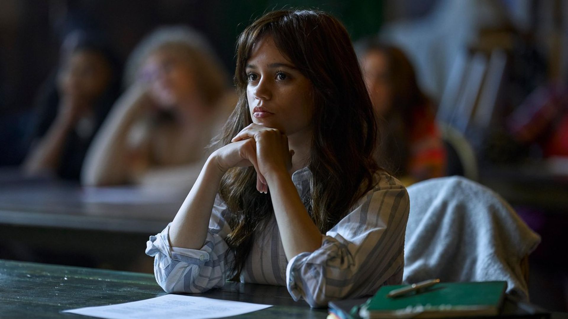 Jenna Ortega con un suéter blanco sentada en un escritorio durante clase con una expresión concentrada