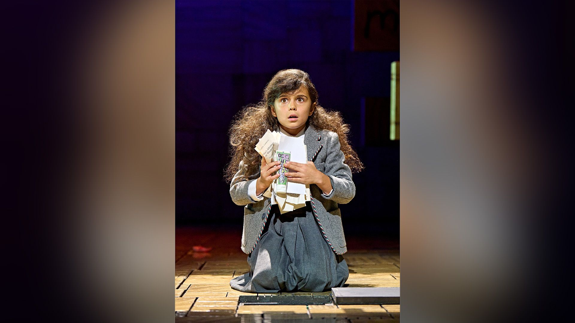 Arabella Stanton en un vestido gris arrodillada en un escenario teatral, sosteniendo hojas de papel arrugadas en las manos