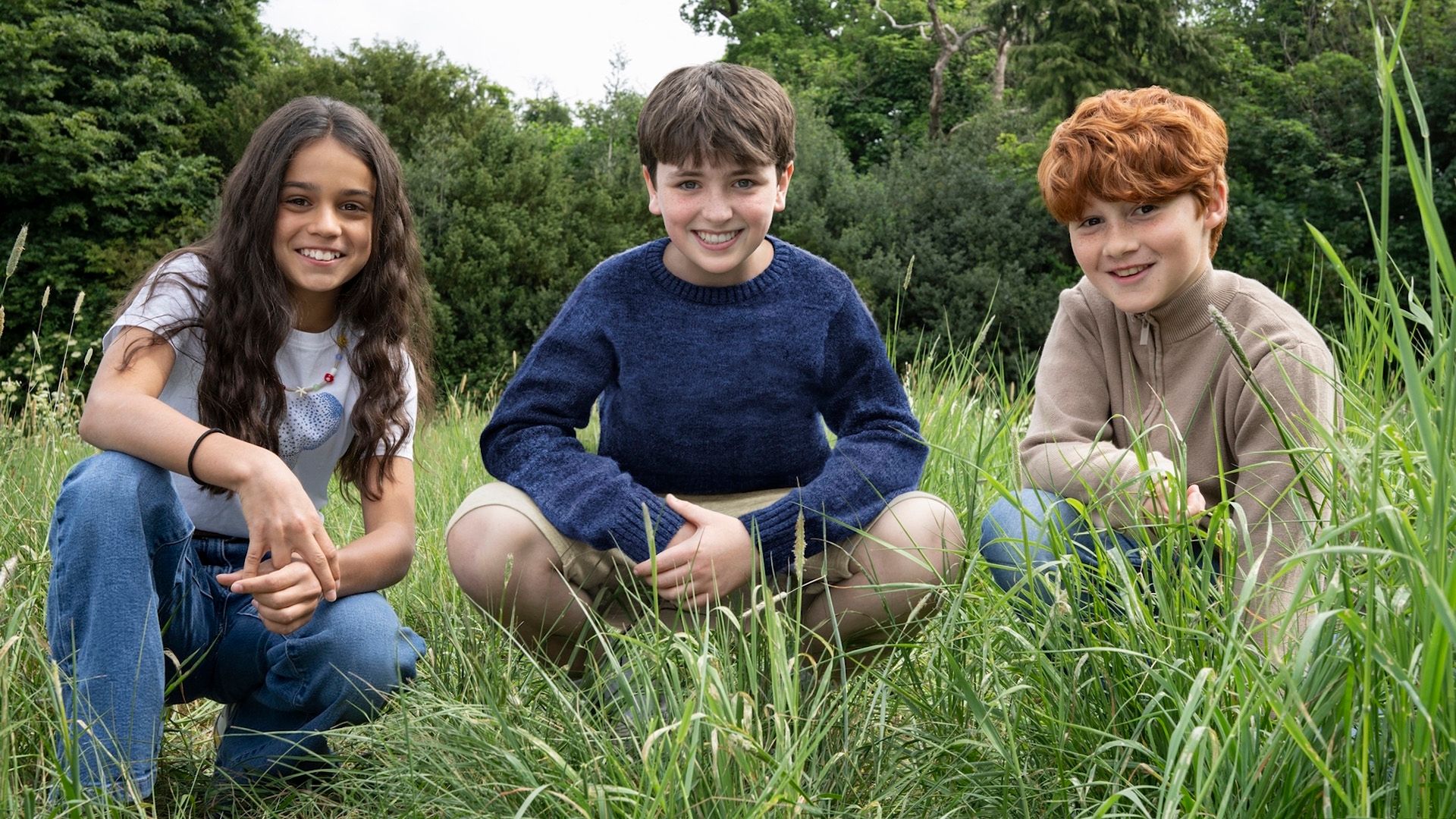 Los jóvenes actores Arabella Stanton, Dominic McLaughlin y Alastair Stout agachados en el césped y sonriendo