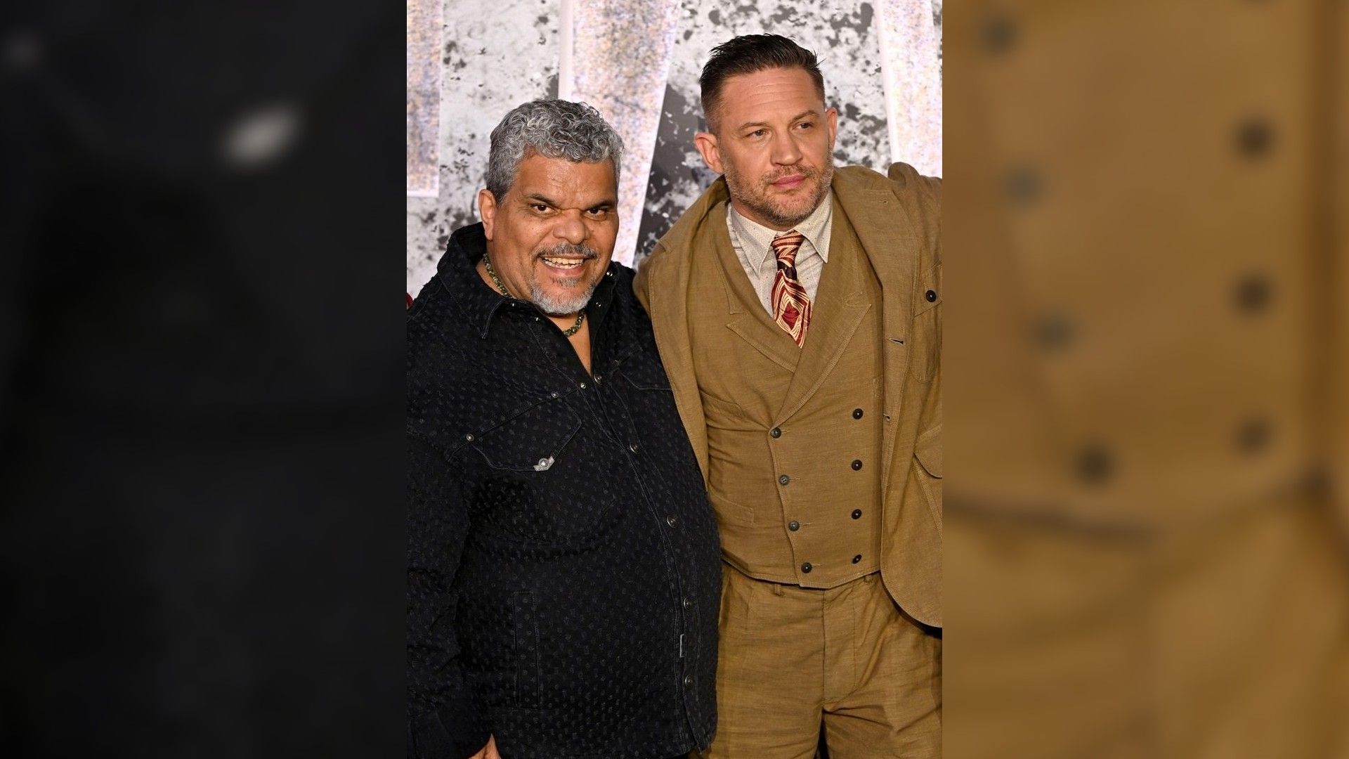 Luis Guzmán y Tom Hardy posan en la alfombra roja: uno con camisa oscura, el otro con traje marrón y corbata