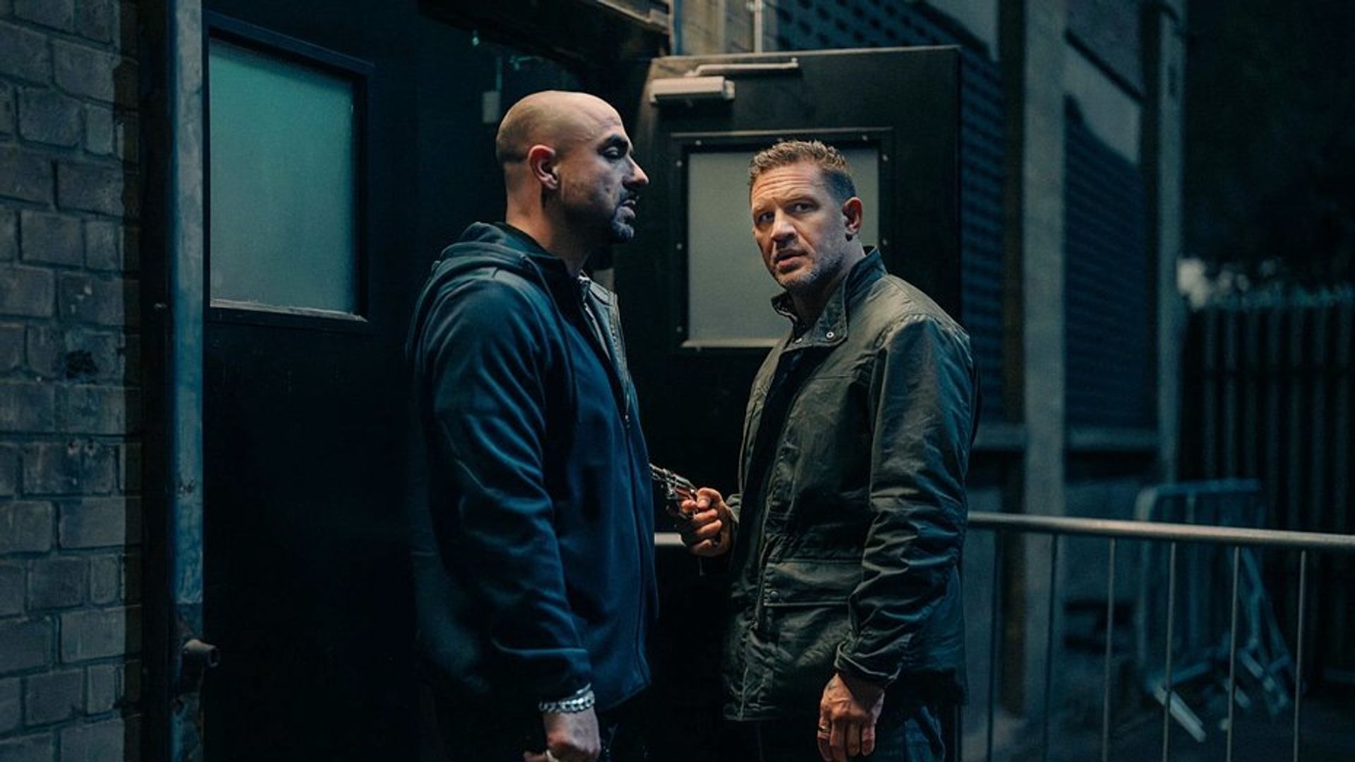 Tom Hardy y otro hombre vestidos de oscuro conversan junto a una salida negra por la noche, uno de ellos sostiene un arma