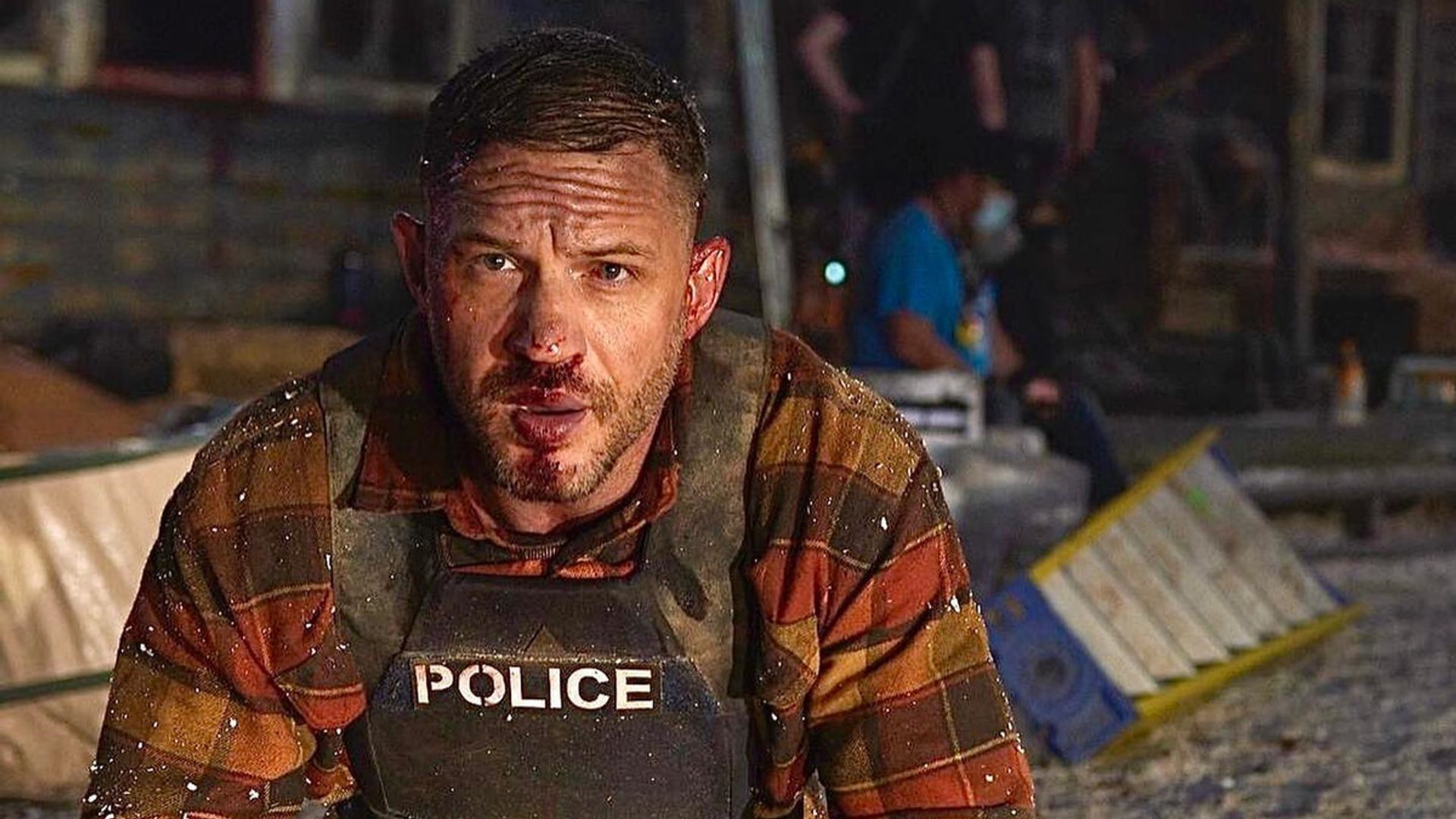 Tom Hardy con chaleco antibalas y rostro golpeado está arrodillado en la escena del incidente