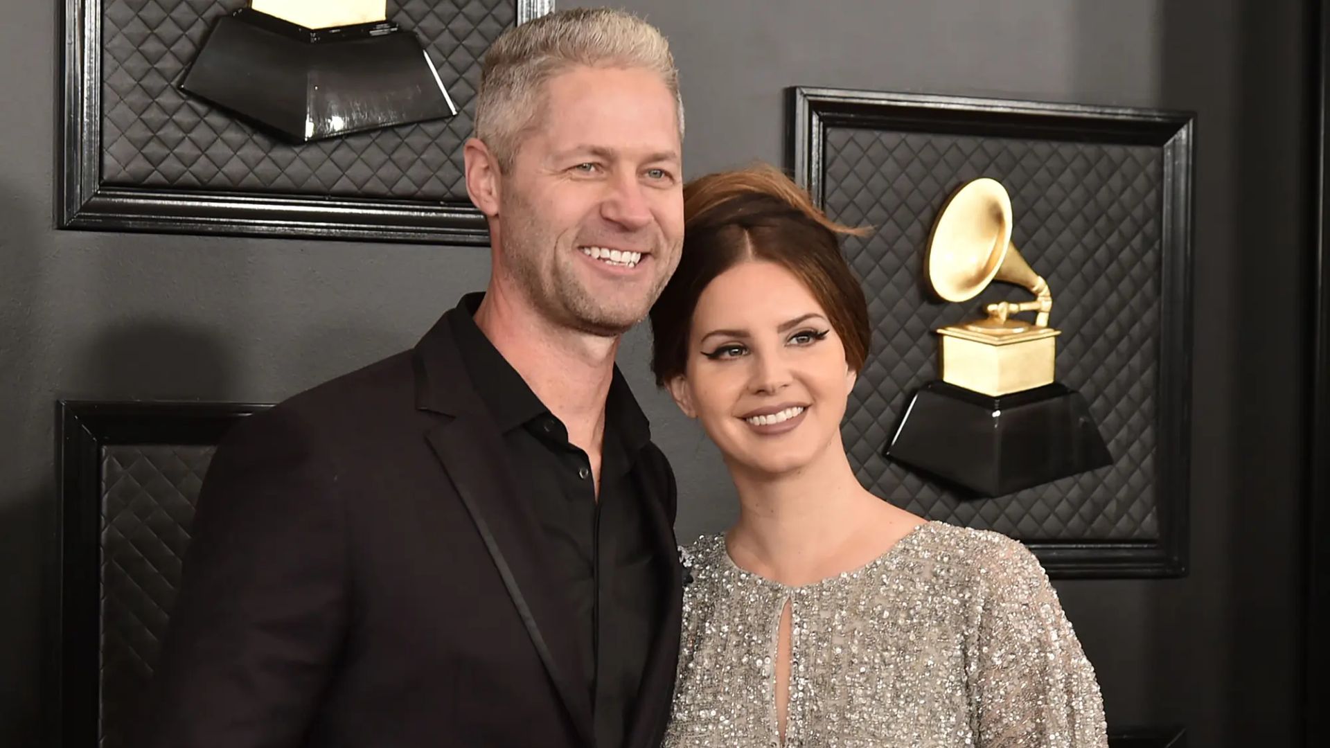 Lana Del Rey y Sean Larkin posan en la alfombra roja de los Grammy Awards contra una pared negra con imágenes de gramófonos dorados