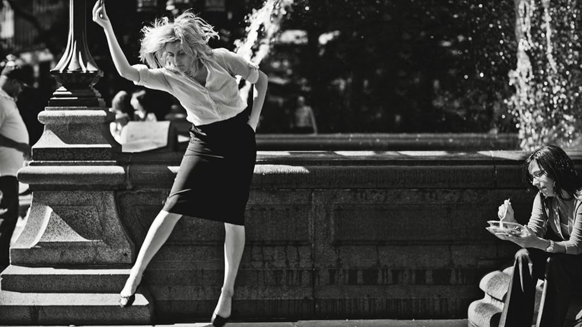 La actriz Greta Gerwig baila con una blusa blanca y falda negra contra el telón de fondo de una fuente