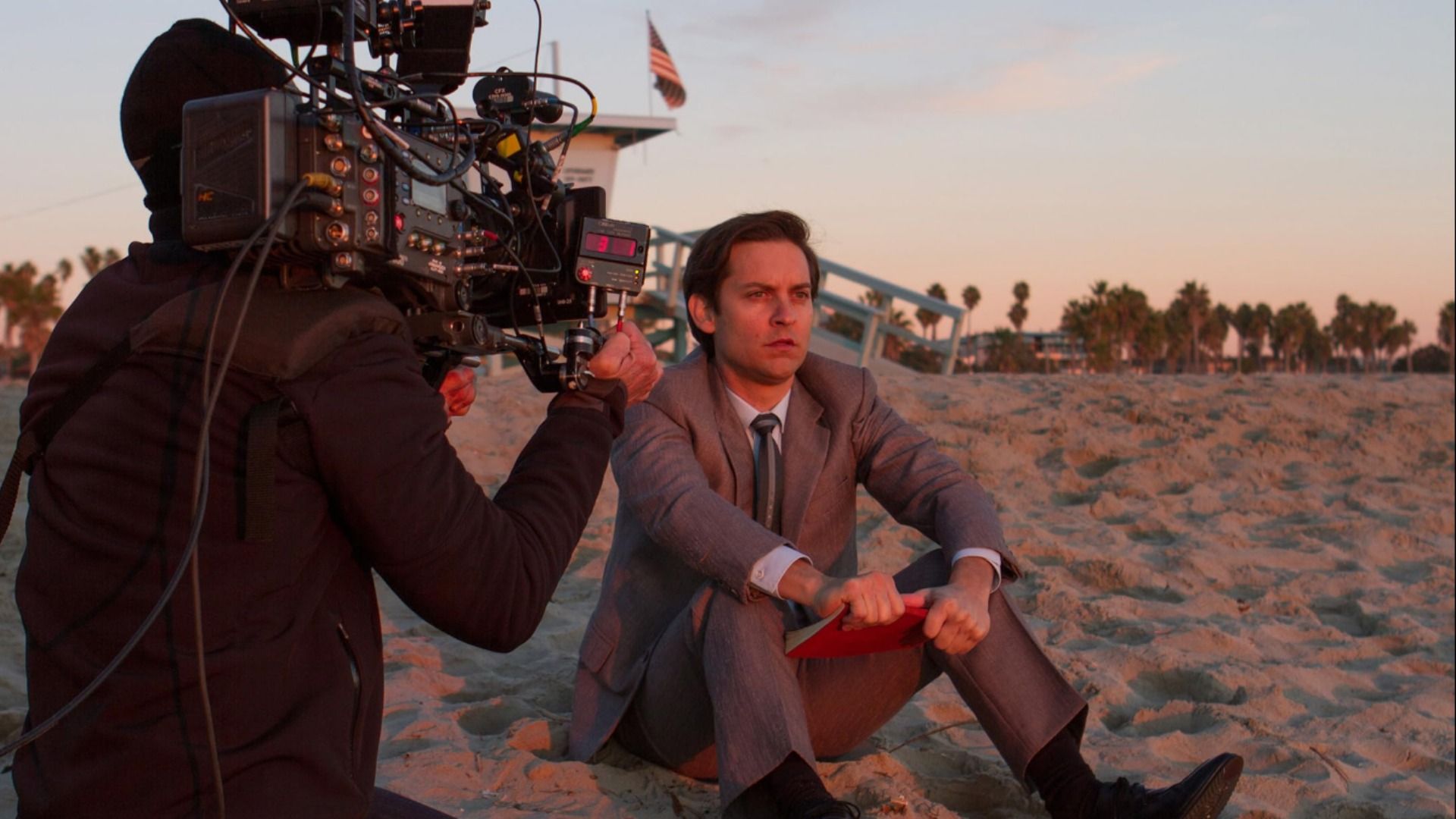 El actor Tobey Maguire con traje gris sentado en la arena