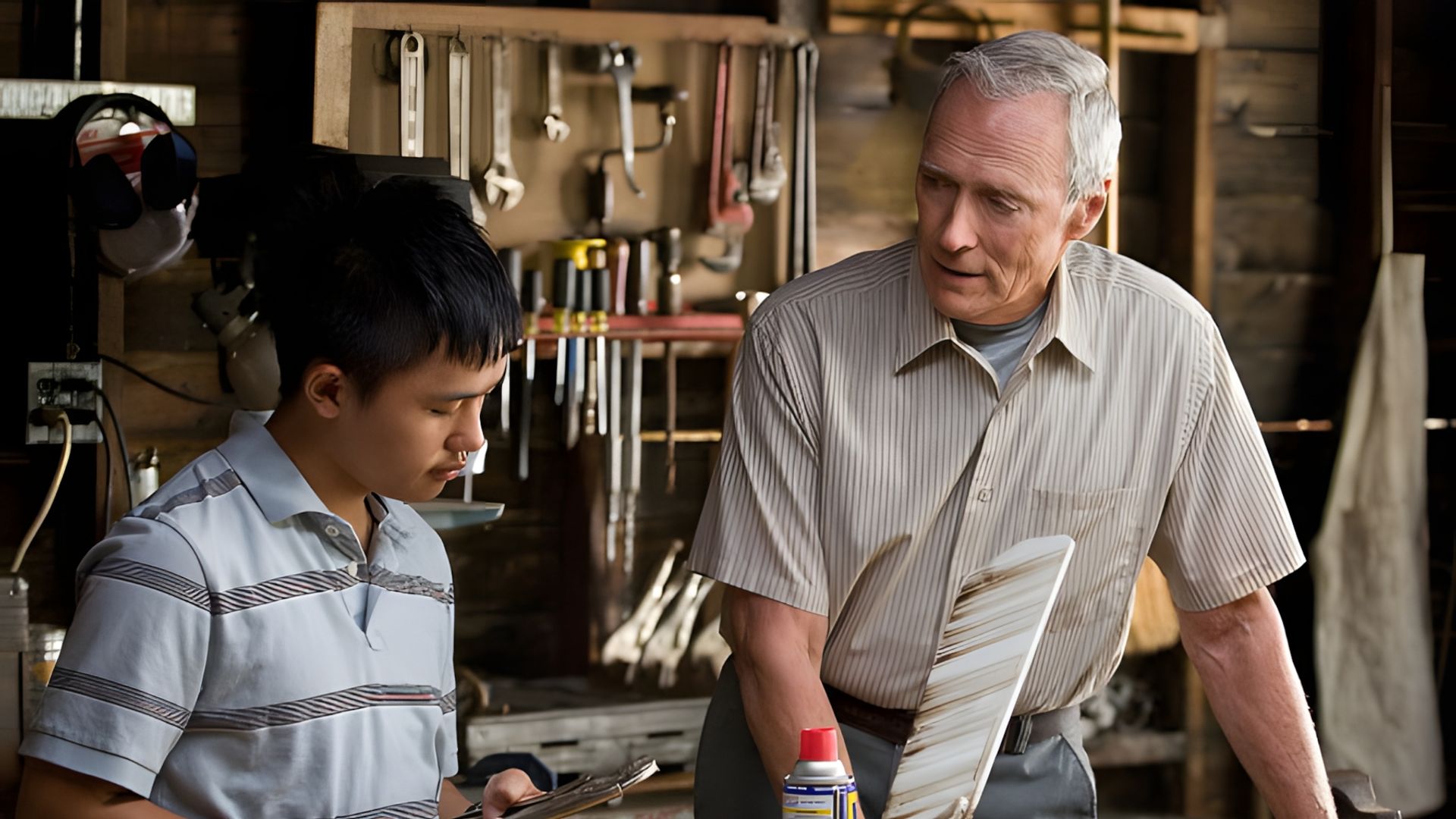 Clint Eastwood y un adolescente trabajan en un taller, discutiendo sobre una herramienta con un banco de trabajo lleno de herramientas colgadas de fondo