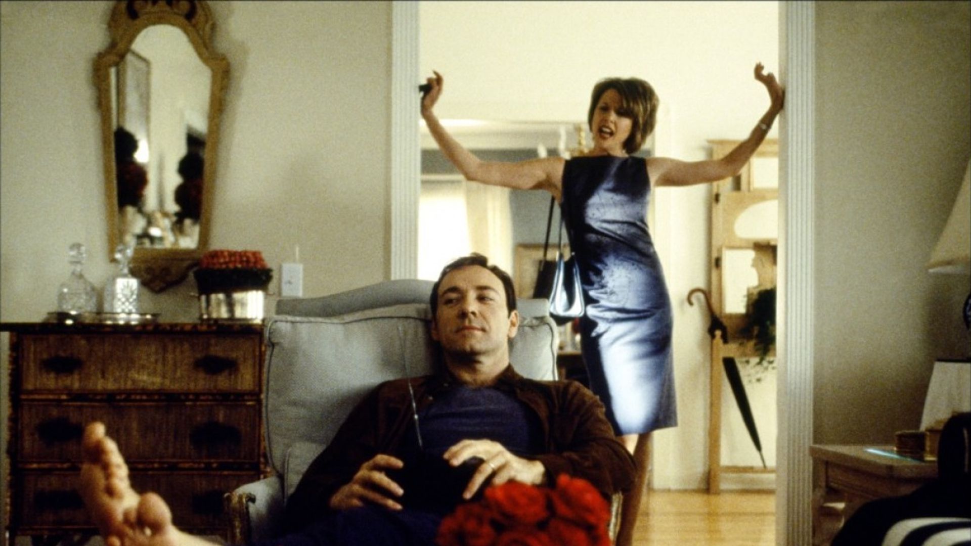 Kevin Spacey sentado relajado en una silla con un control remoto mientras una mujer con vestido de noche detrás de él gesticula emocionalmente en la puerta