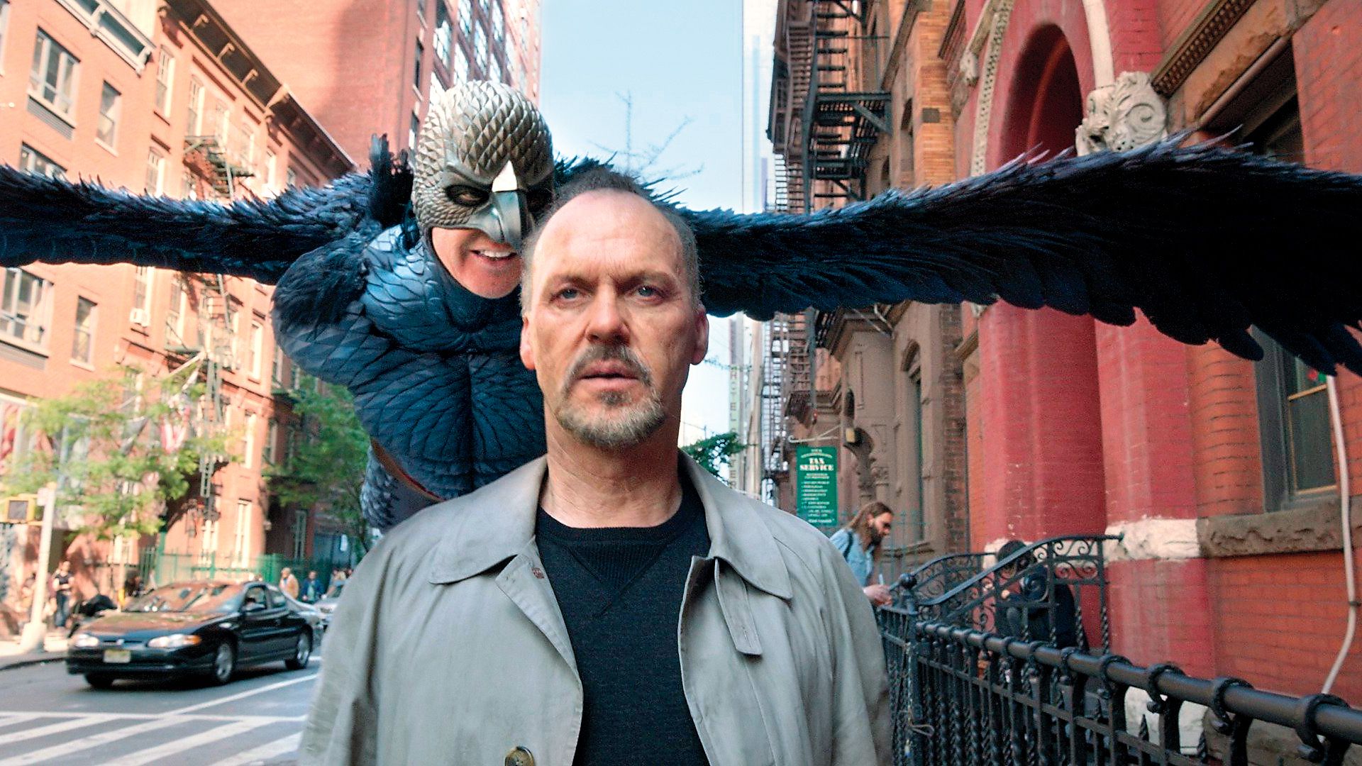 Michael Keaton camina por la calle mientras detrás de él flota un personaje fantástico con alas y máscara de pájaro