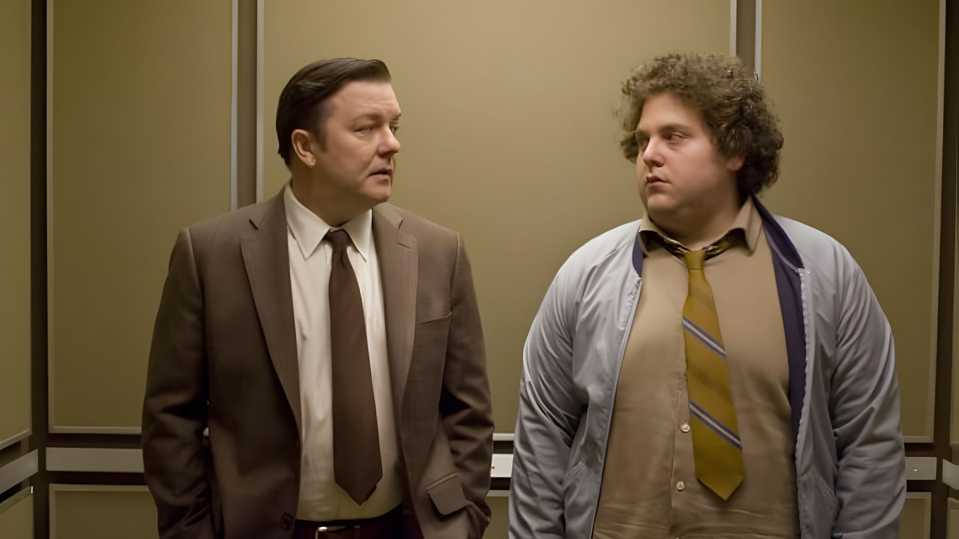 Ricky Gervais y Jonah Hill vestidos de traje de pie en un ascensor, mirándose con expresiones serias