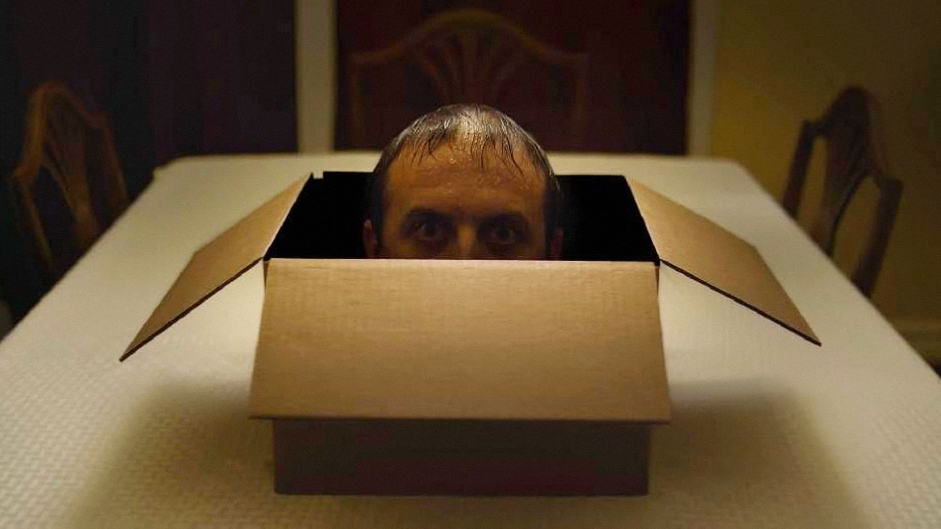 El rostro de un hombre se asoma desde una caja marrón sobre una mesa