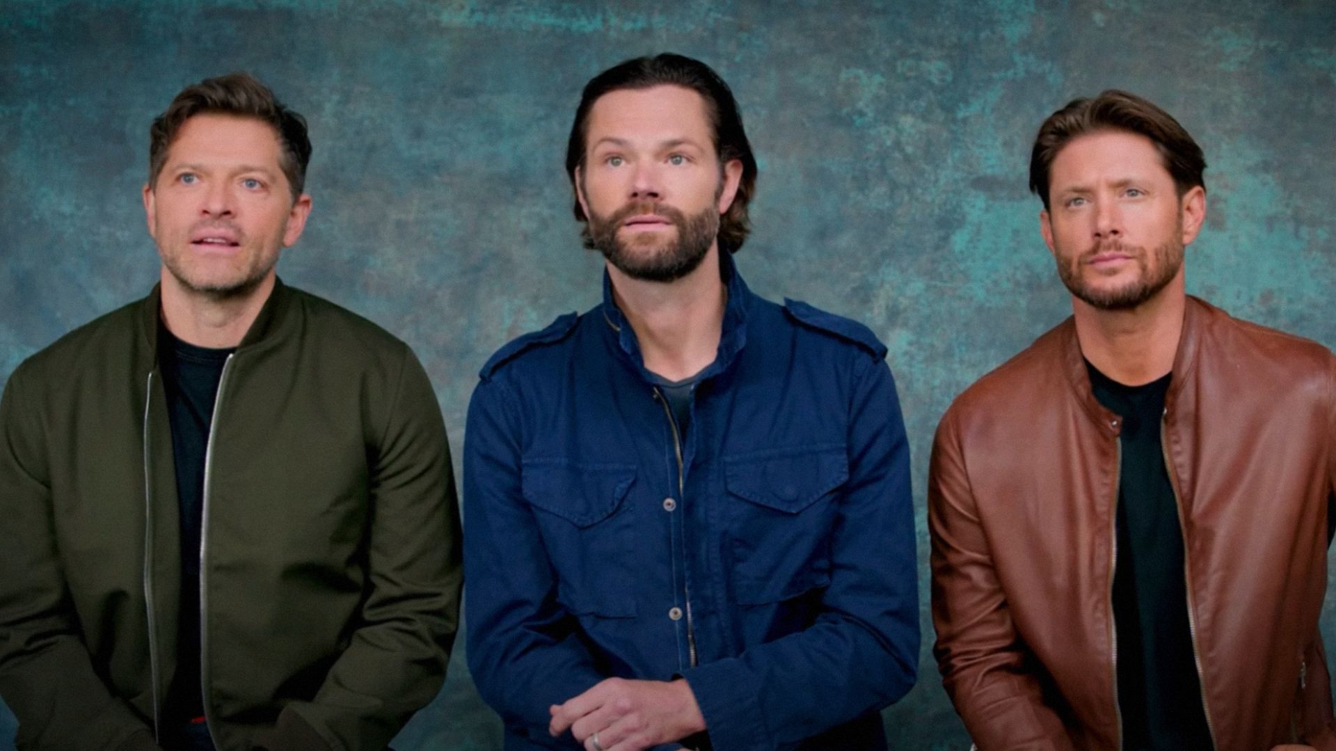 Los actores Misha Collins, Jared Padalecki y Jensen Ackles sentados contra un fondo azul llevando chaquetas coloridas