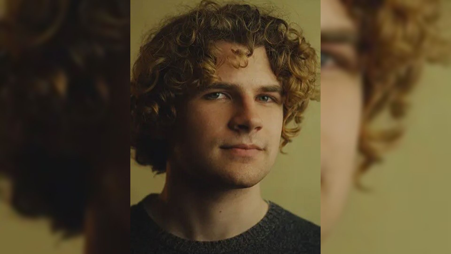 Un joven de cabello rizado en un suéter negro parado contra un fondo amarillo